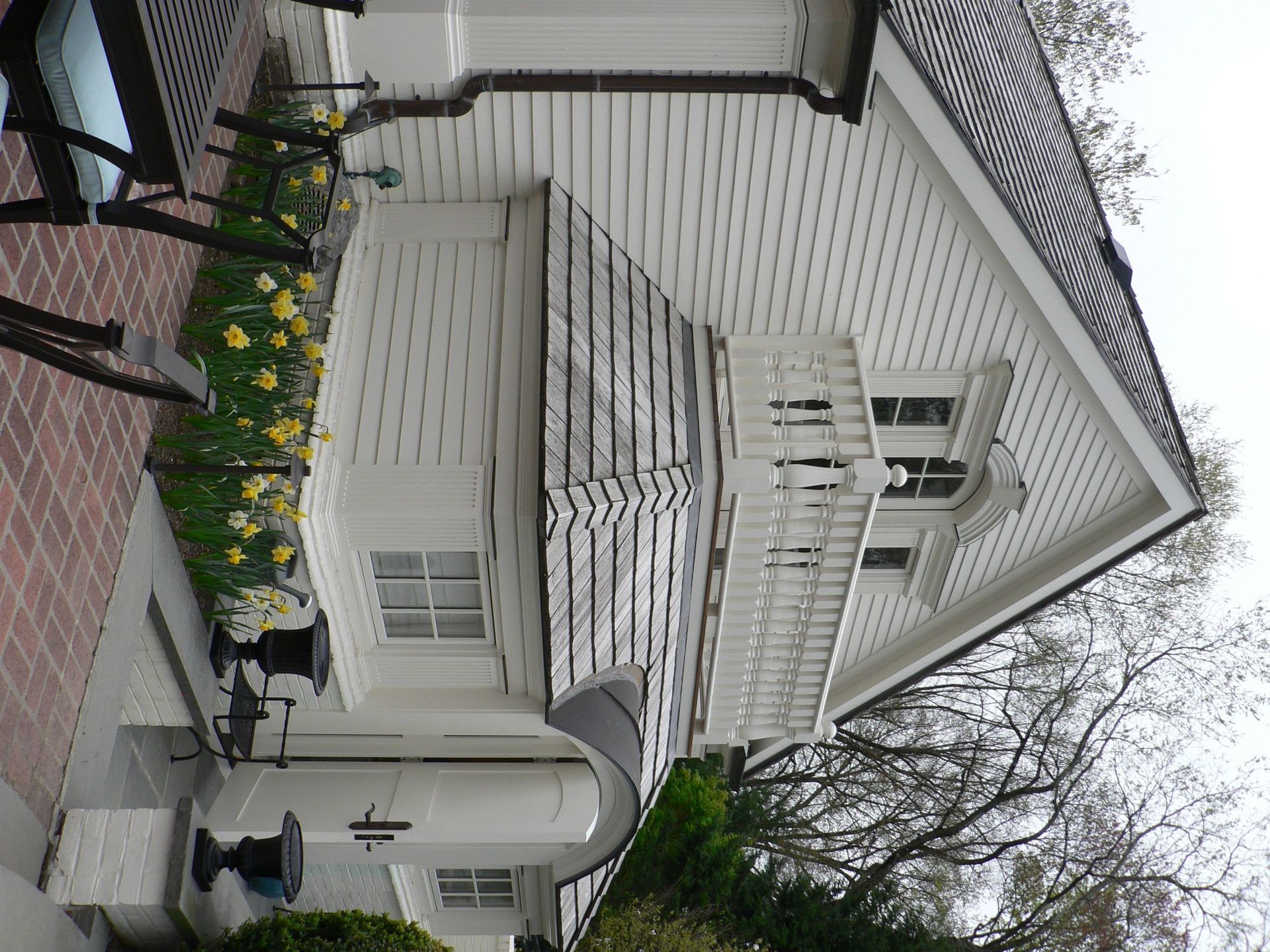 White house with black roof, brick patio, and a bench with yellow flowers.