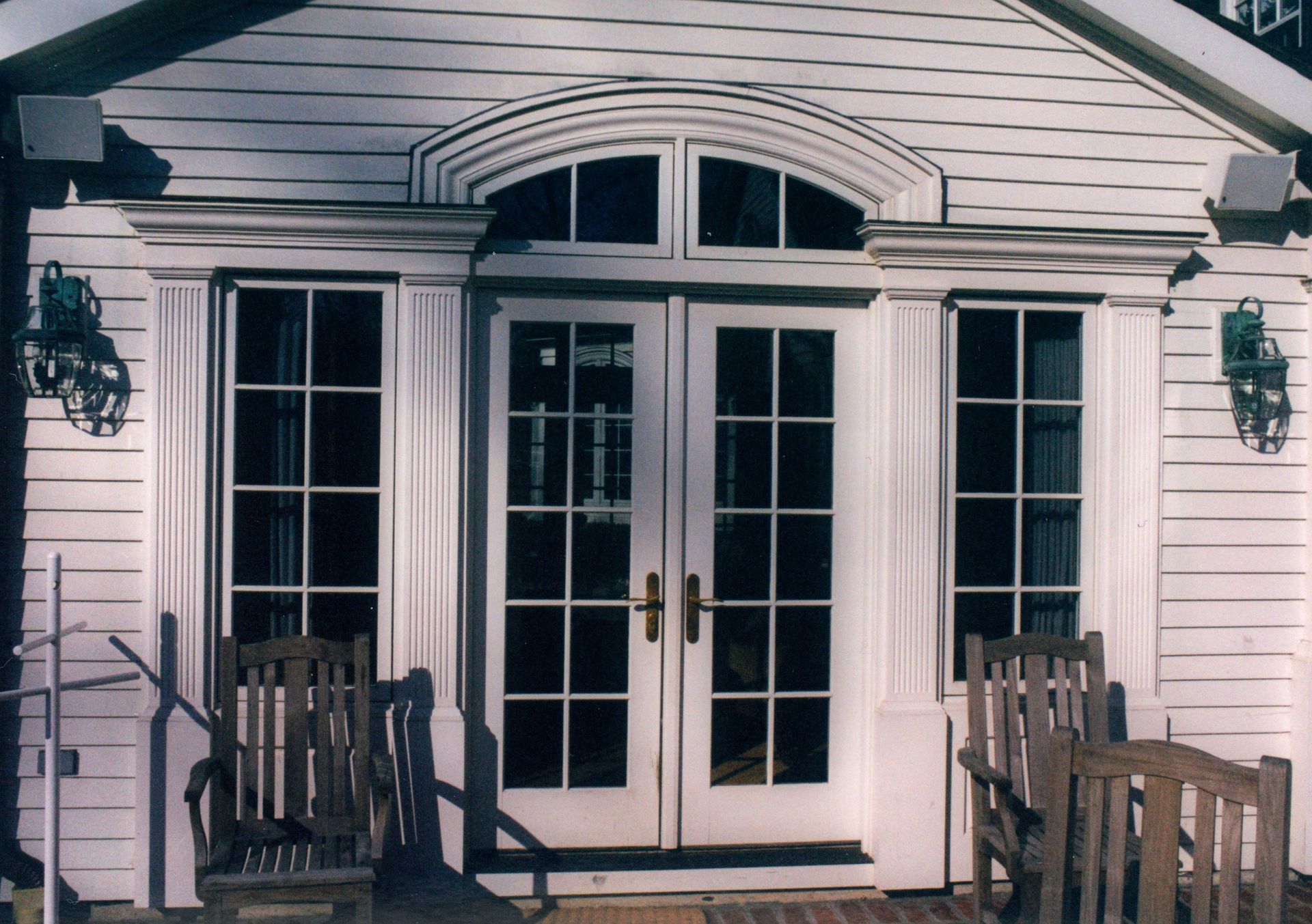 White exterior with arched window and French doors. Two chairs on a porch.
