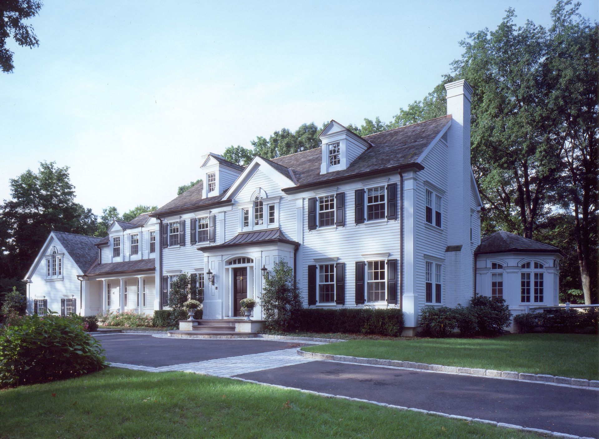 White two-story house with dark shutters, front door, and long driveway, surrounded by trees and green lawn.