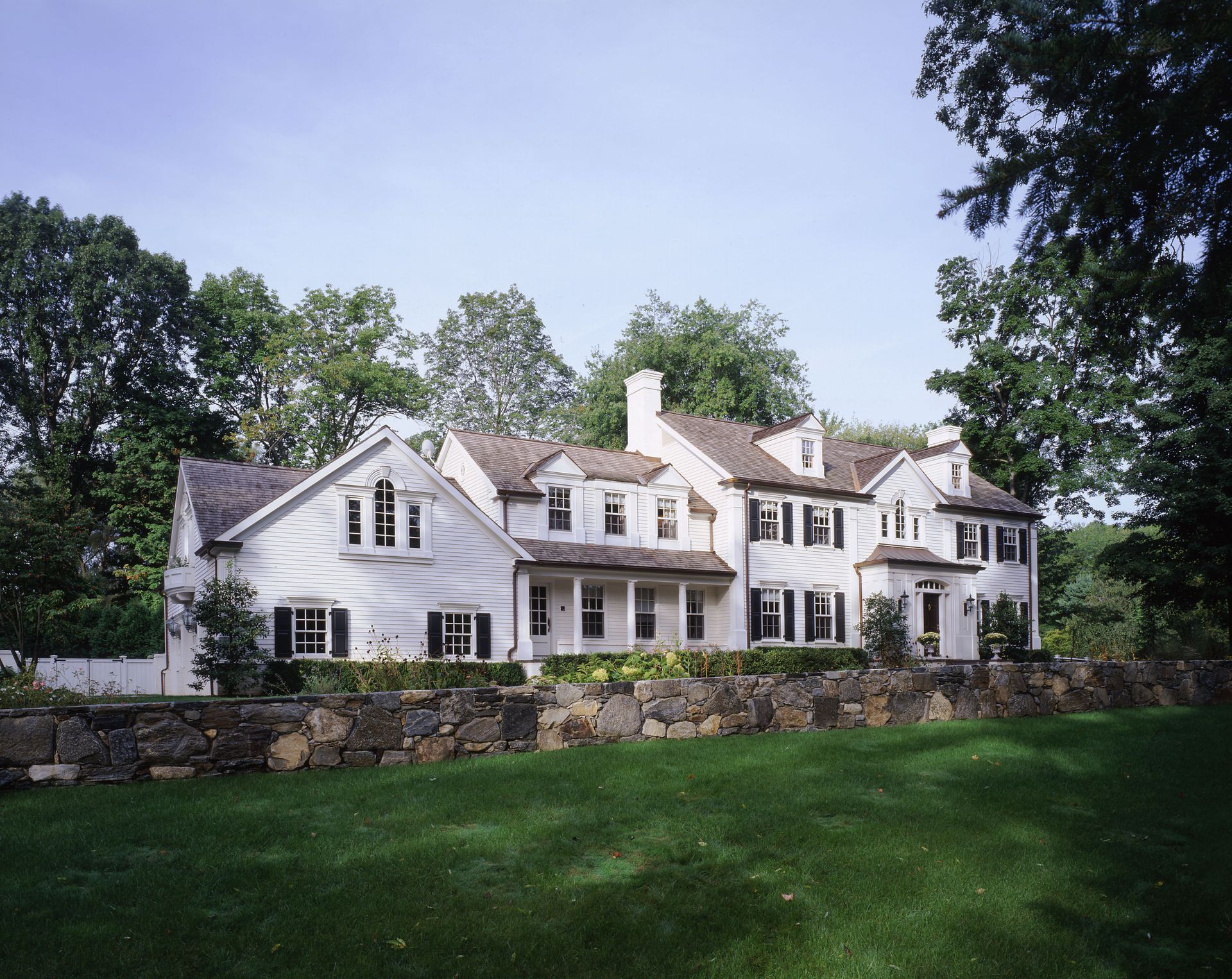 White house with black shutters, on a green lawn, behind a stone wall. Trees frame the property.