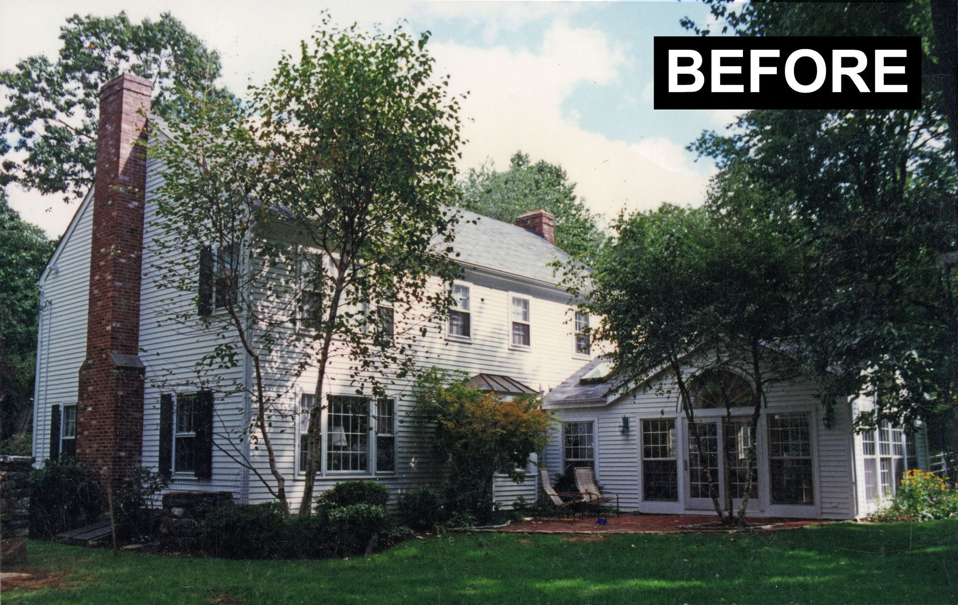 White two-story house with brick chimney, surrounded by trees. 