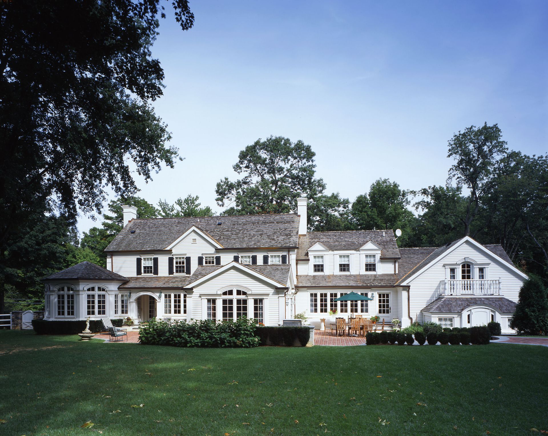 White two-story house with shingled roof, windows, and a large lawn, surrounded by trees.