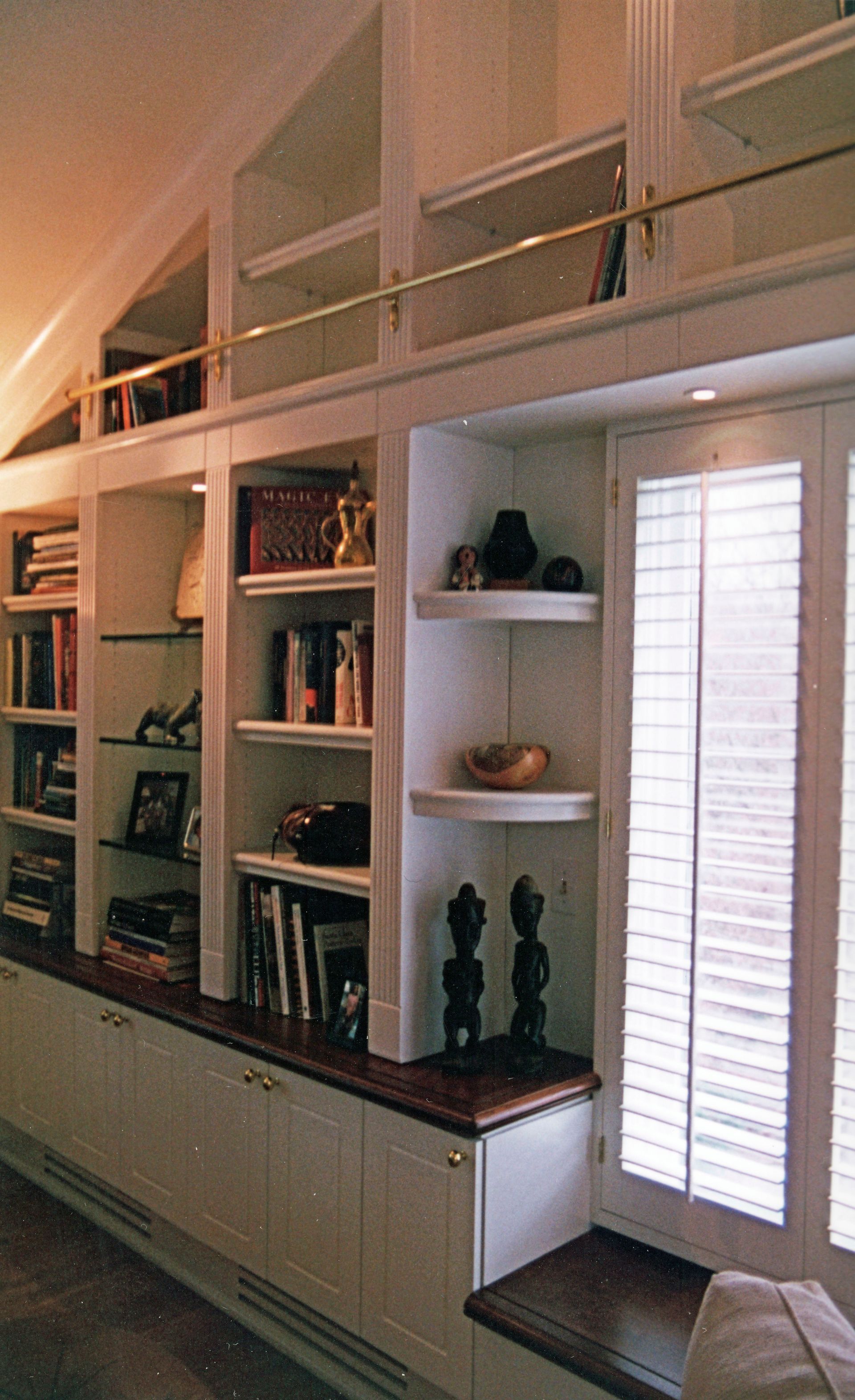 Built-in white bookshelves with books and decor, brass rail, and window with shutters.
