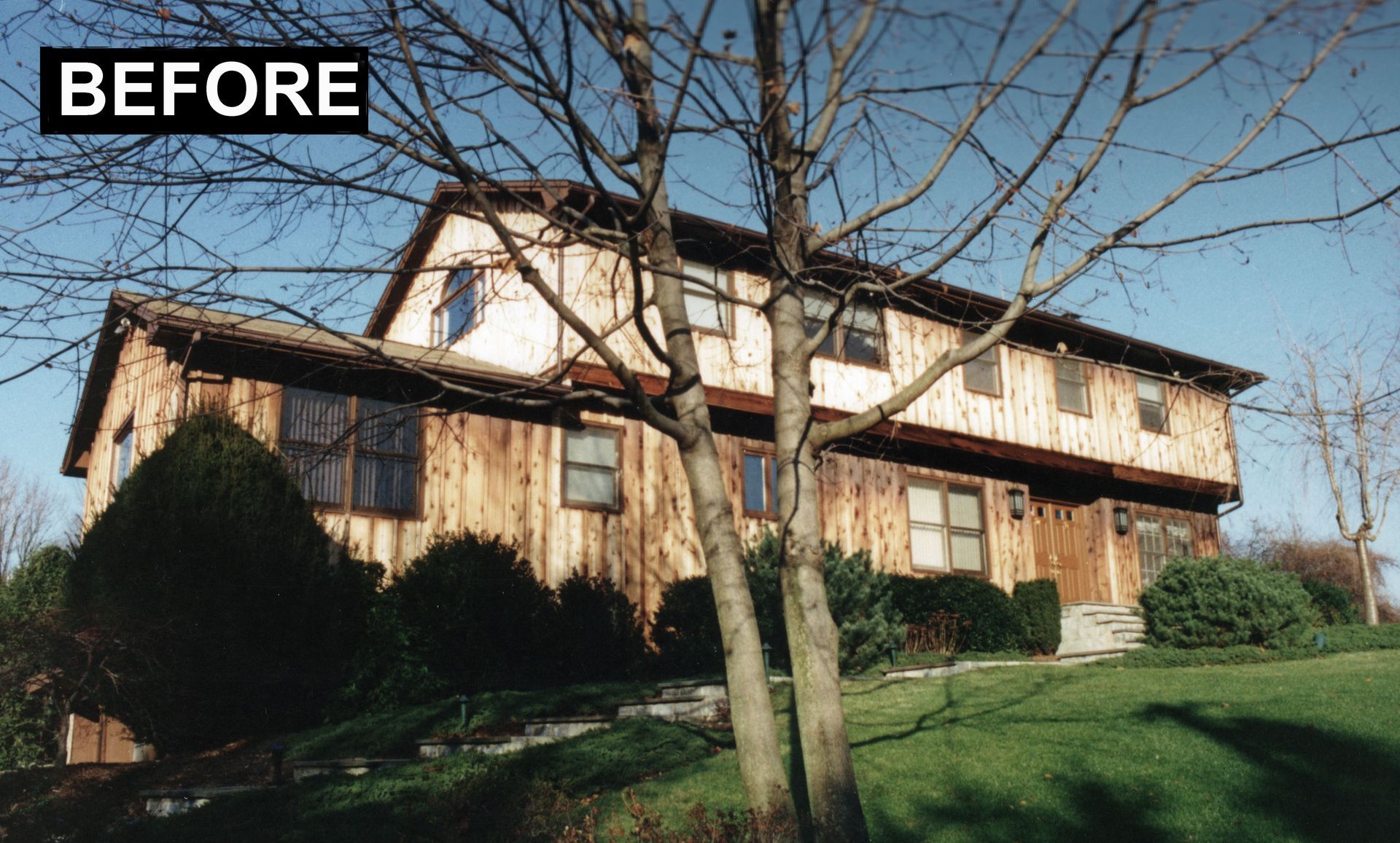 Two-story wooden house with brown siding, shrubs, and a bare tree. 