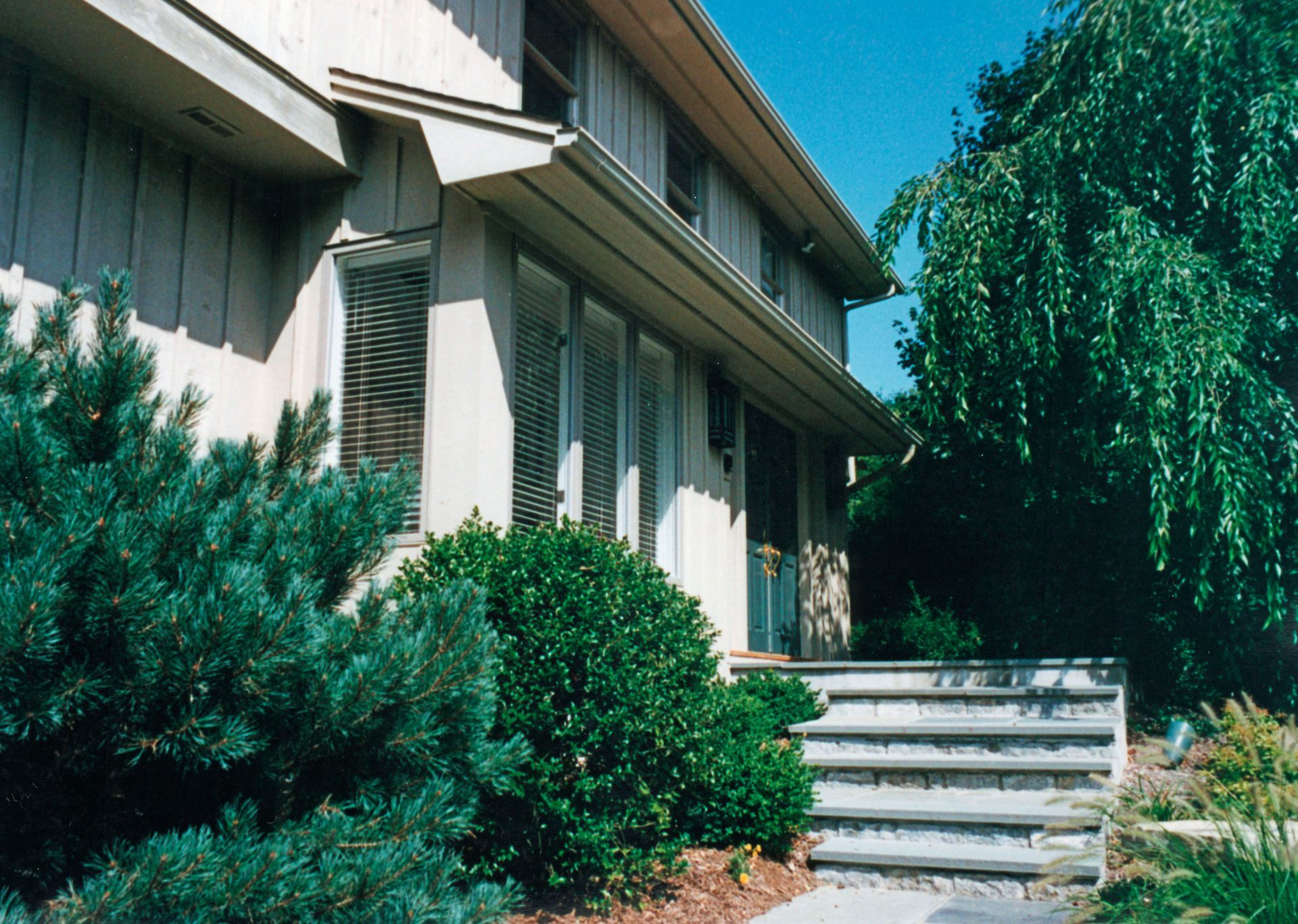 Two-story house with tan siding, steps, and greenery.