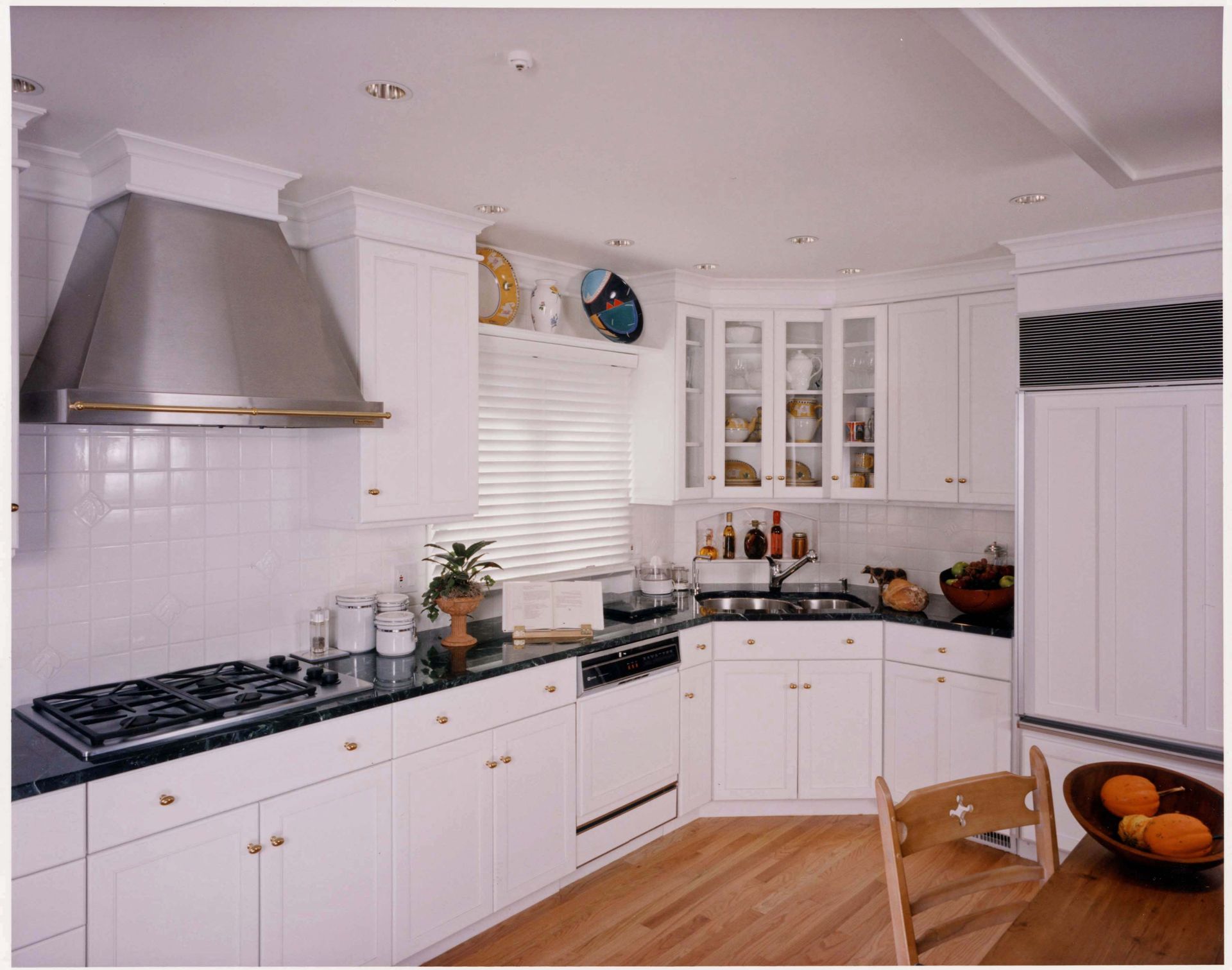 White kitchen with stainless steel hood, white cabinets, dark countertops, and wooden floors.