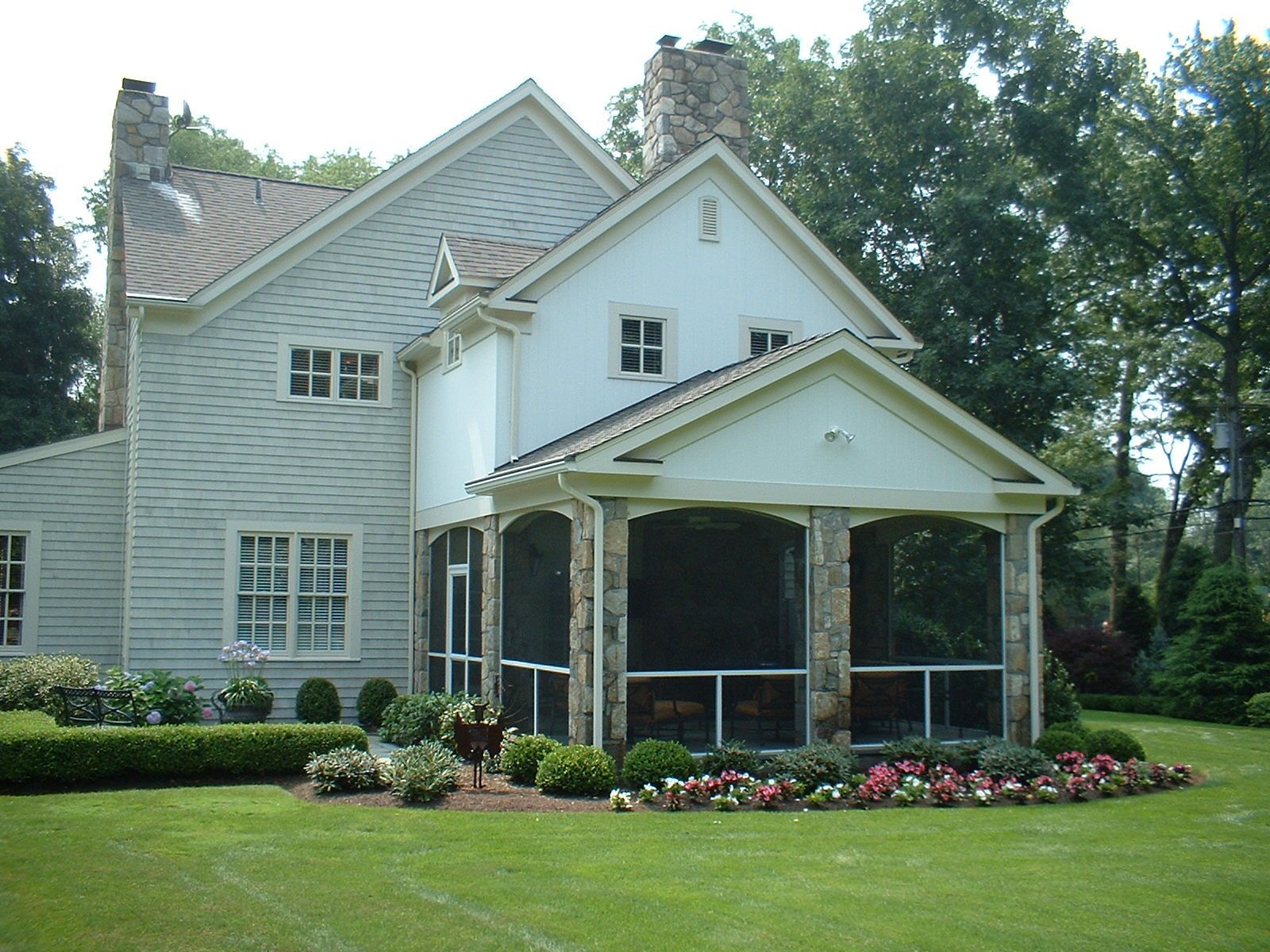 Exterior of a two-story light gray house with a stone-columned screened porch, surrounded by green lawn and landscaping.