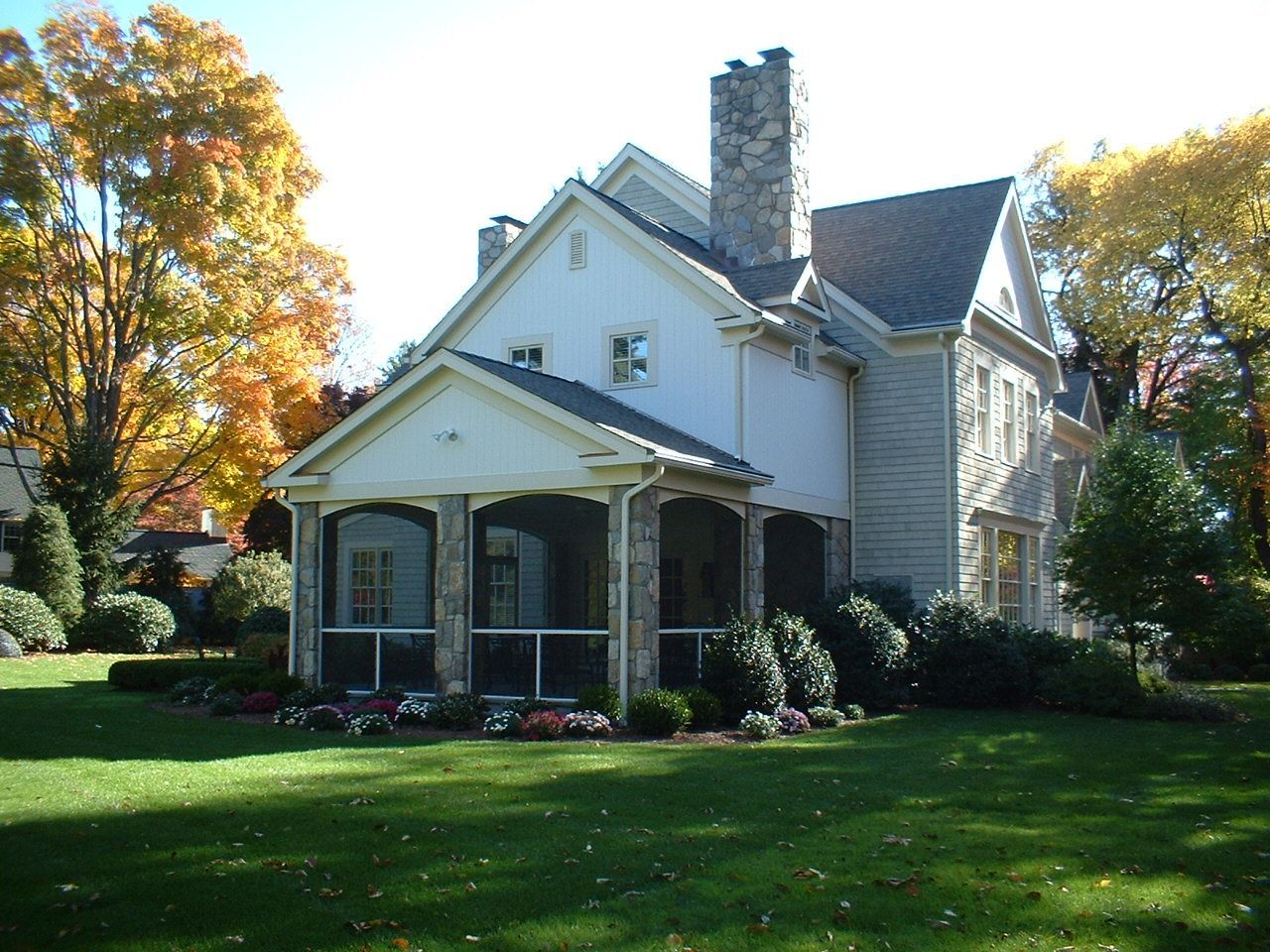 Two-story house with screened porch, stone columns, and manicured lawn surrounded by trees with fall foliage.