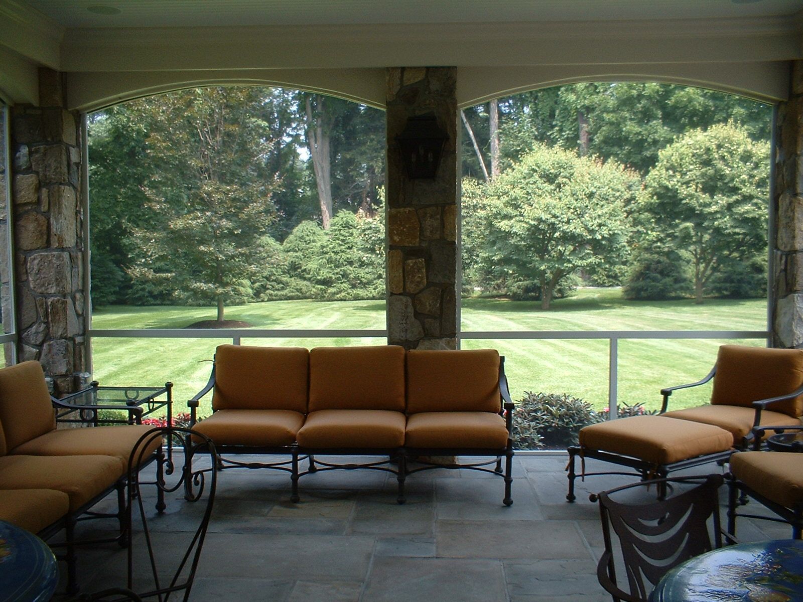 Outdoor screened-in patio with orange cushioned furniture, stone columns, and a view of a grassy yard with trees.