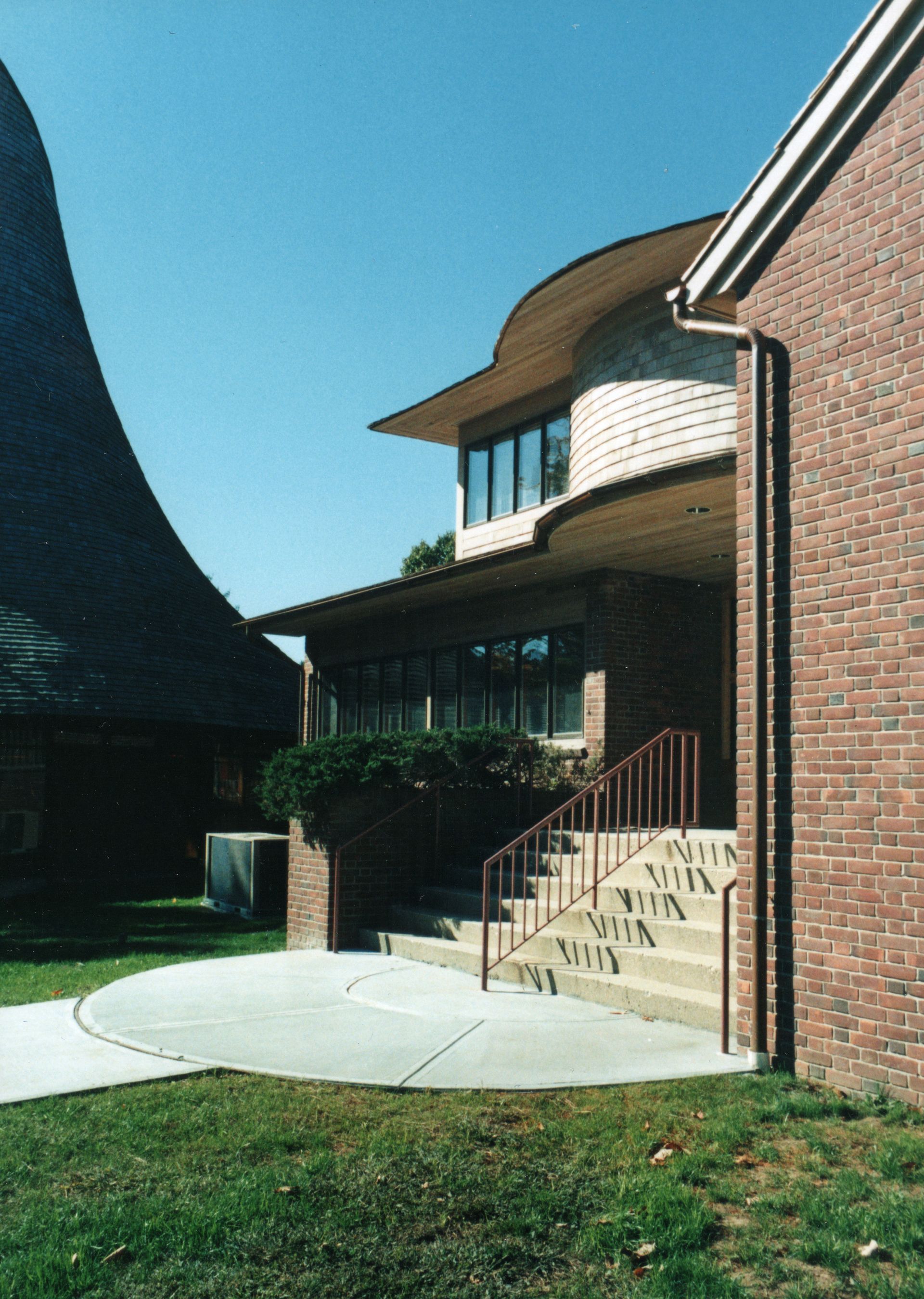 Brick building with curved roof extension; steps lead to entrance with bushes.