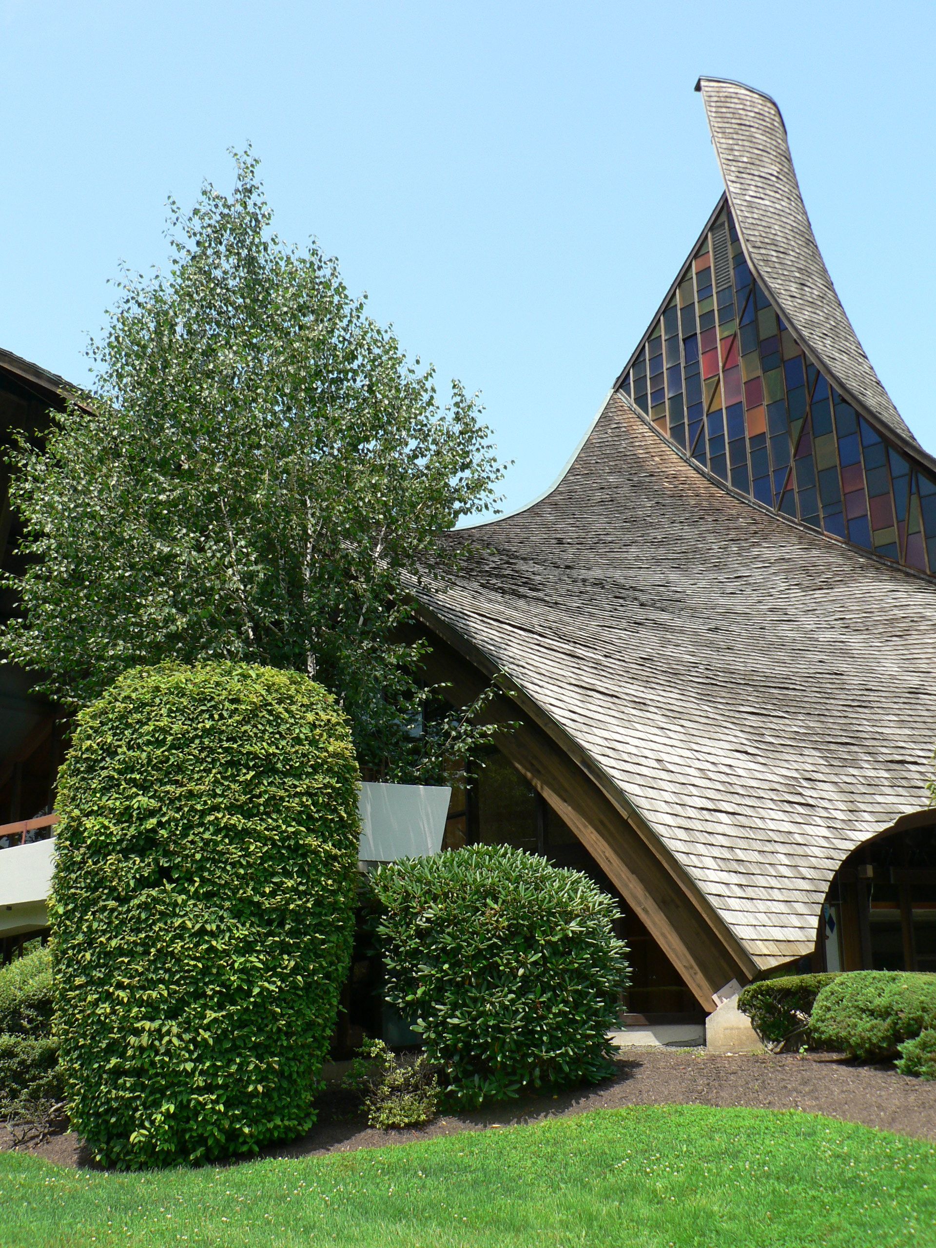 A modern building with a curved, textured roof against a clear blue sky. Green shrubs and trees in foreground.