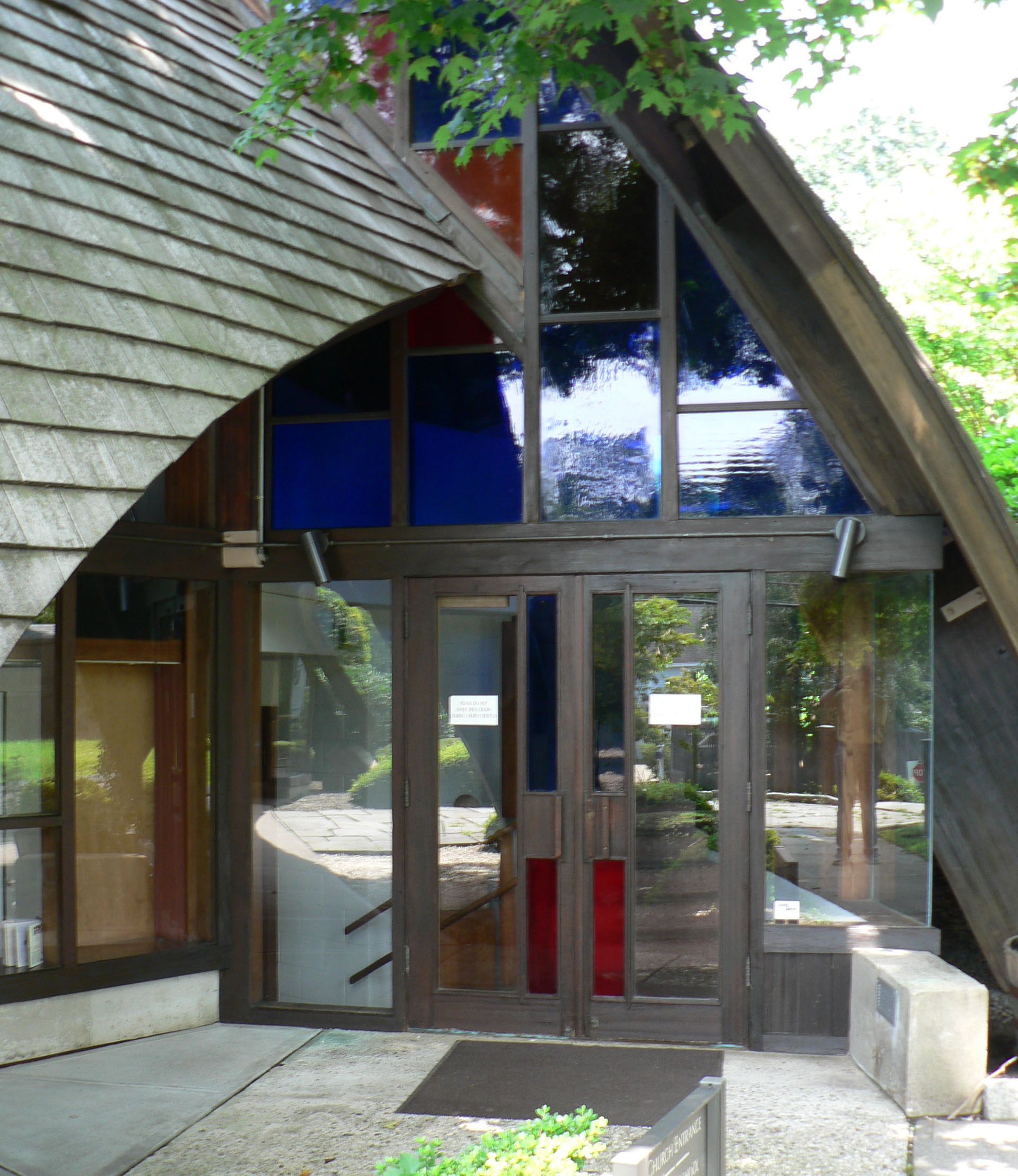 Entrance to a building with a wood shingled roof and stained-glass windows, some blue and red.