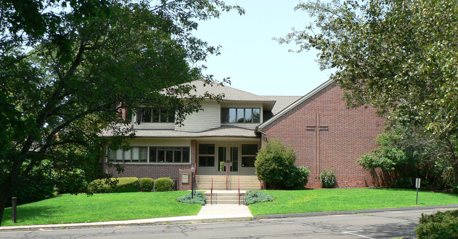 Two-story building with a light-colored front facade, red brick side, green lawn, and trees.