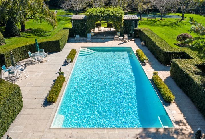 Rectangular pool surrounded by stone patio, hedges, and lawn, with lounge chairs and a pergola.