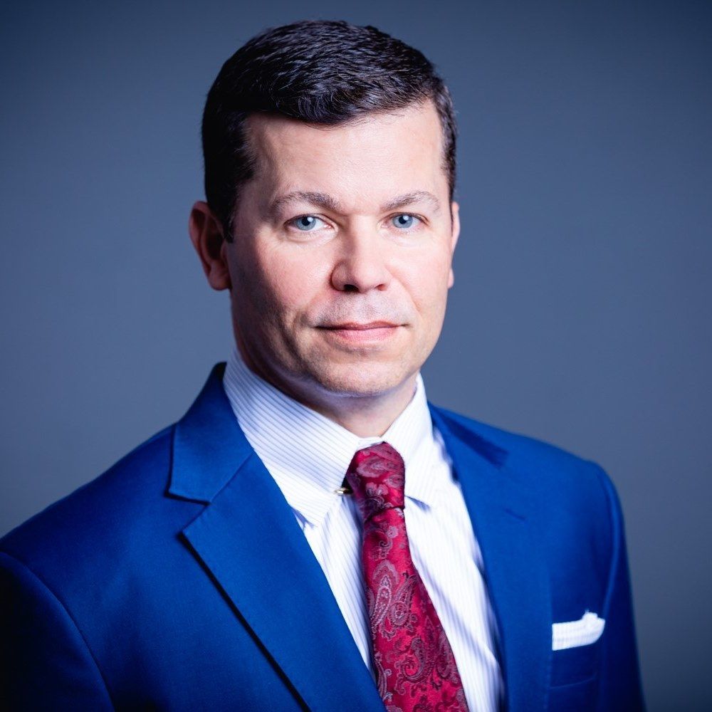 Man in blue suit, red tie, white shirt, looking forward against a gray background.