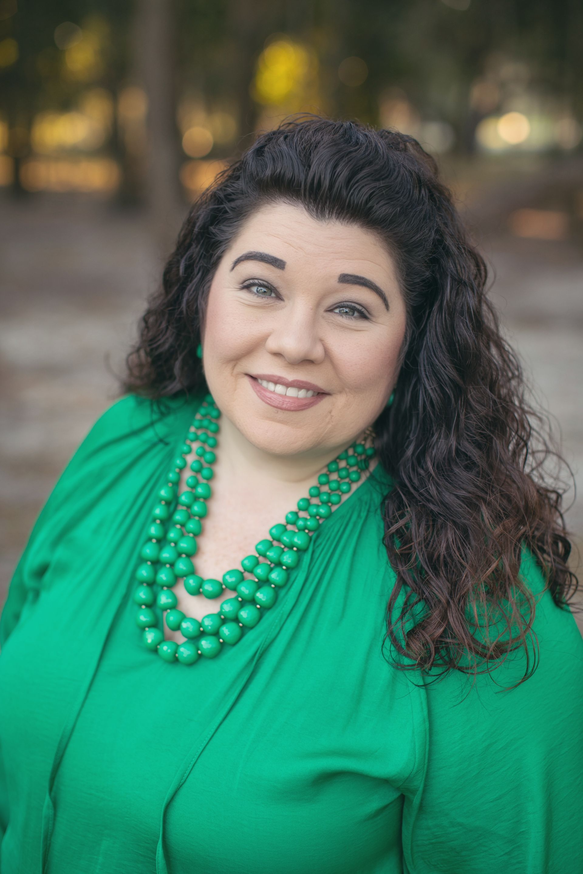 Woman with curly hair smiles, wearing a green blouse and beaded necklace, outdoors.