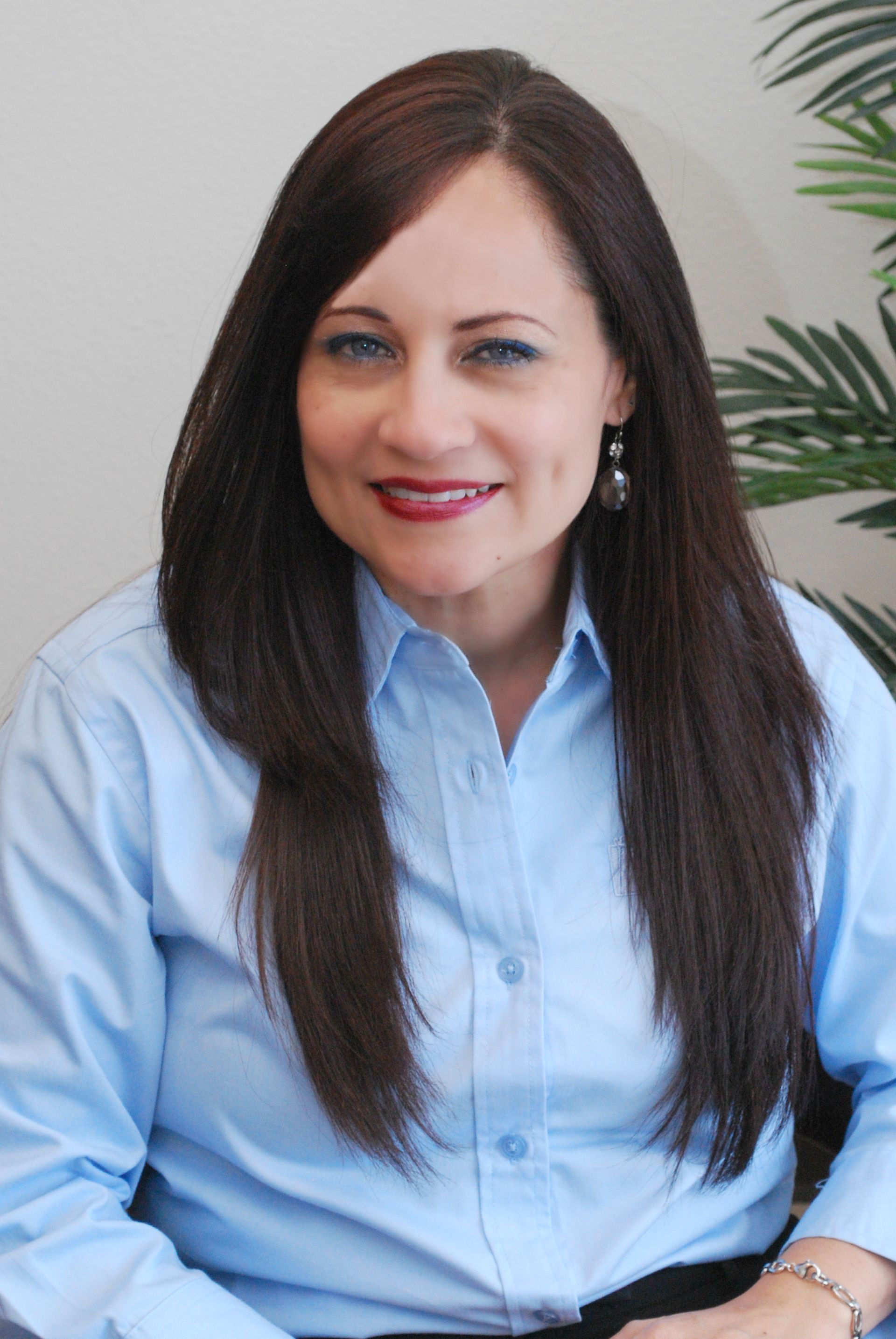 Woman in blue button-down shirt smiling, long dark hair, wearing earrings, near a plant.