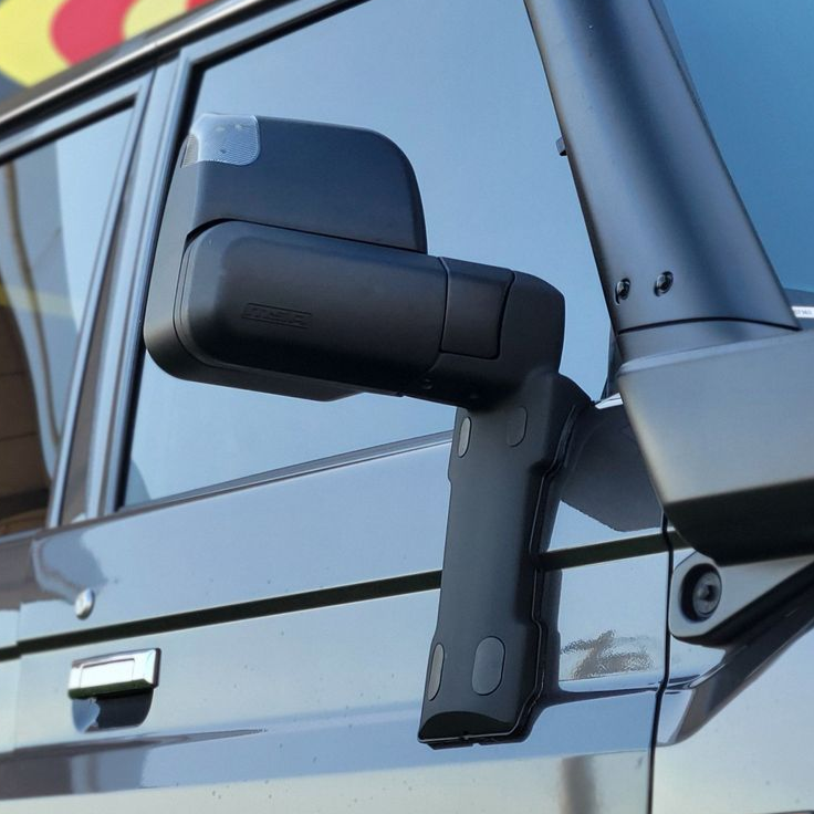 Close-up of a white truck's side mirror, extended for towing a camper trailer on a grassy field.