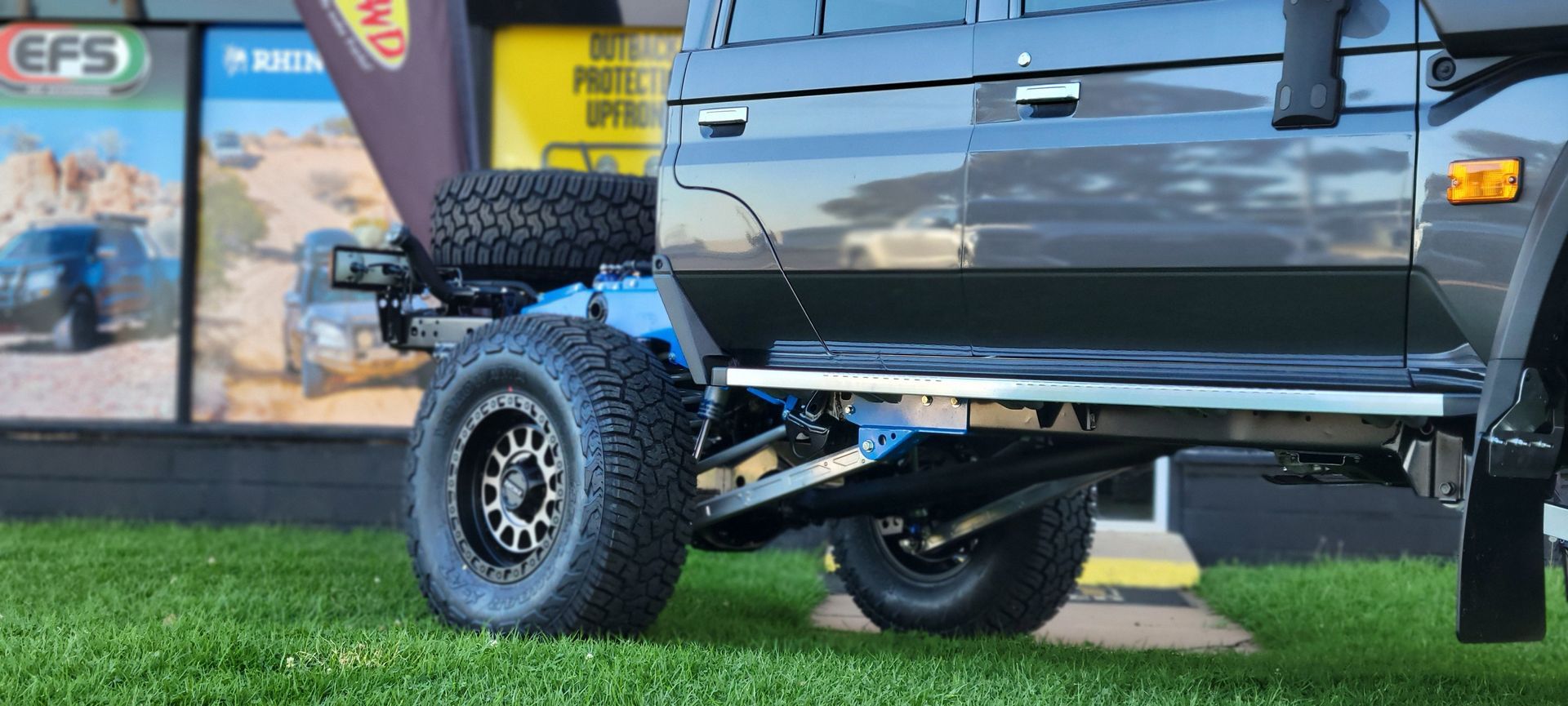 Blue off-road SUV angled down a dirt hill, muddy tires, bright blue sky, green grass and scrub.