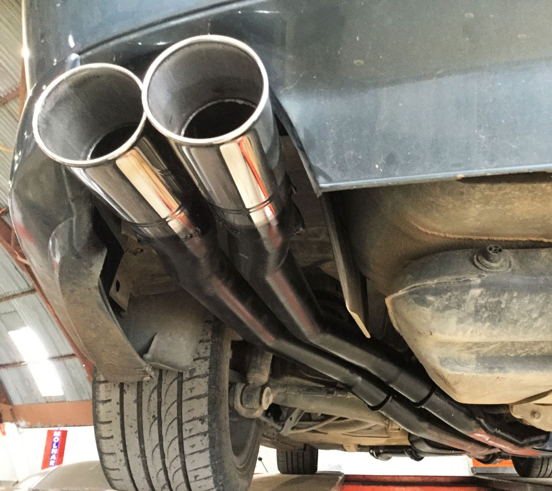 Mechanic working under a lifted truck in a garage. Blue lift, concrete floor, white car nearby.