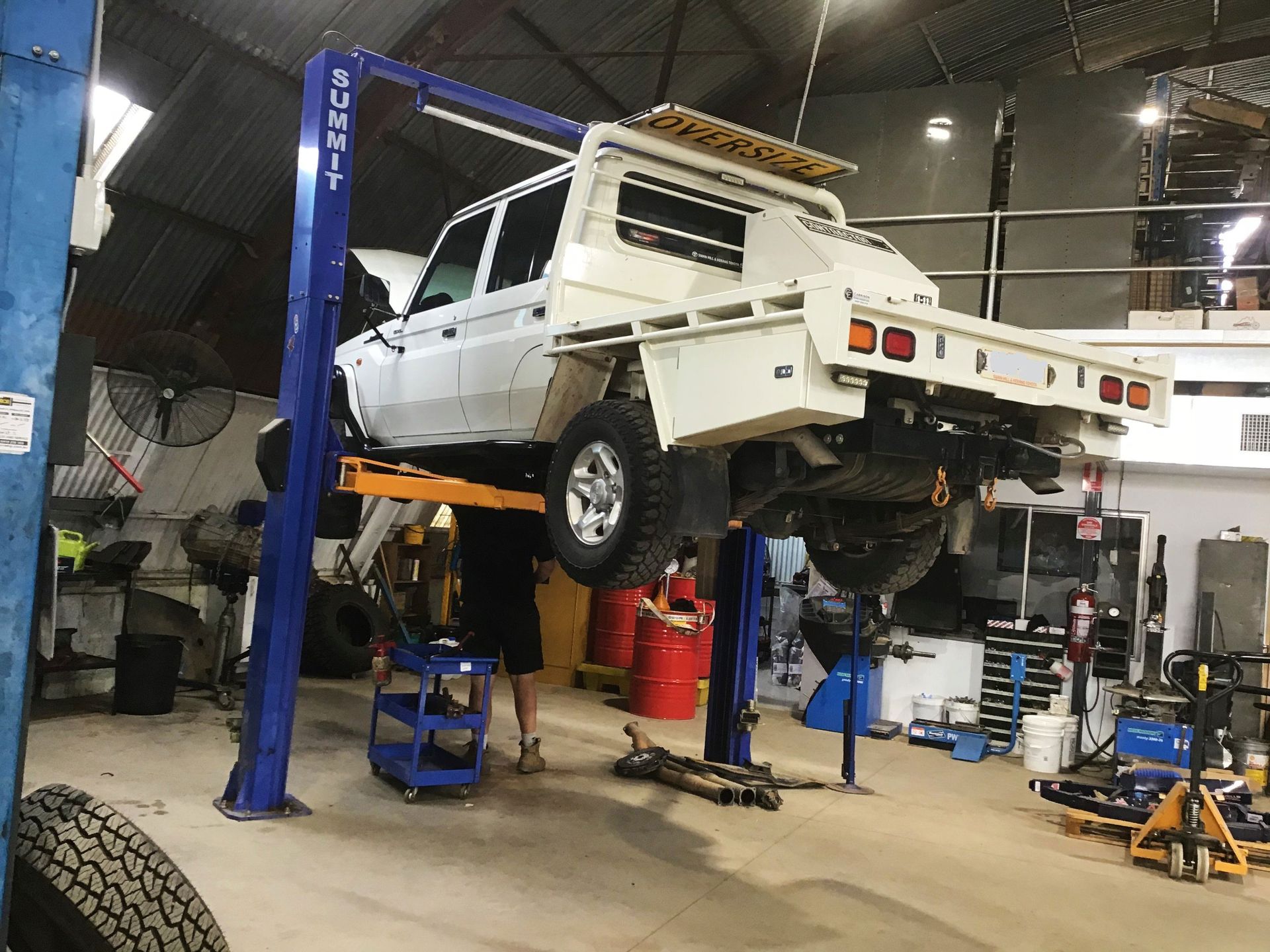 A white truck is lifted on a blue car lift in a workshop; a mechanic works underneath.