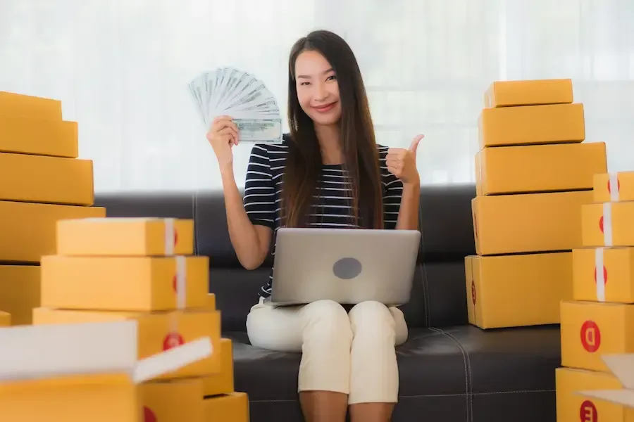 Successful e-commerce woman holding a stack of money with shipping boxes from her online store.