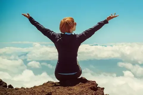 Lady sitting on a mountain above the clouds with her arms in the air. She is on top of the world.