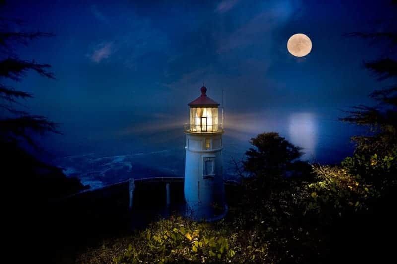 A lighthouse is lit up at night with a full moon in the background.