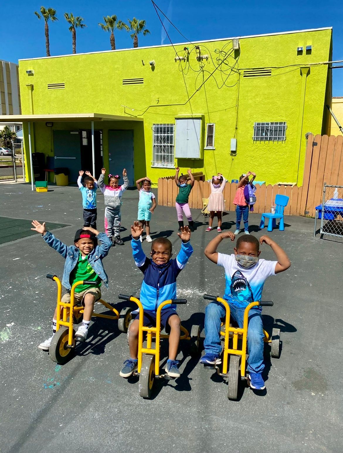 Children on tricycles and standing, raising arms in a playground with a lime-green building in the background.