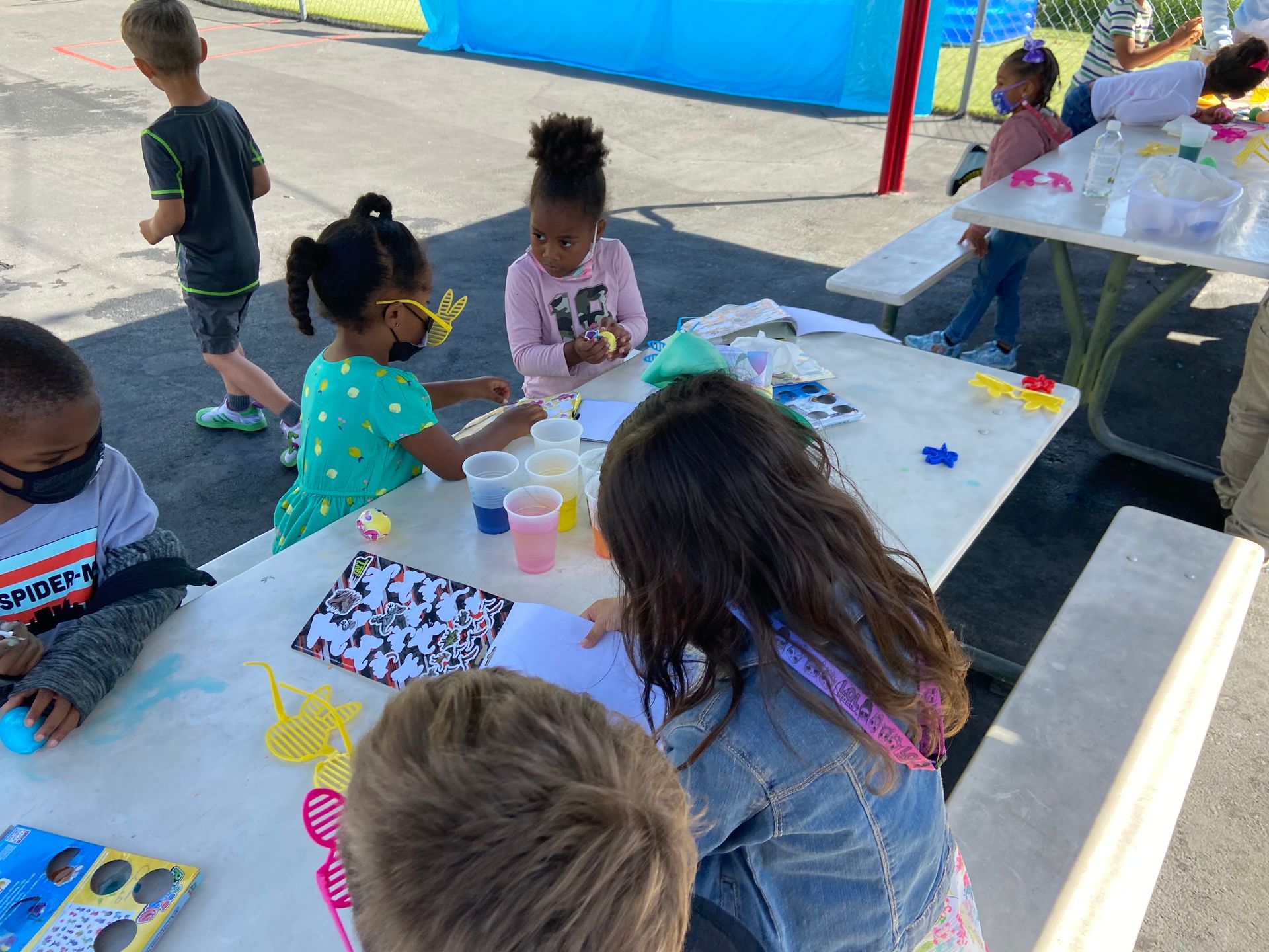 Children at an outdoor table doing a craft, using colorful liquids and cutouts.