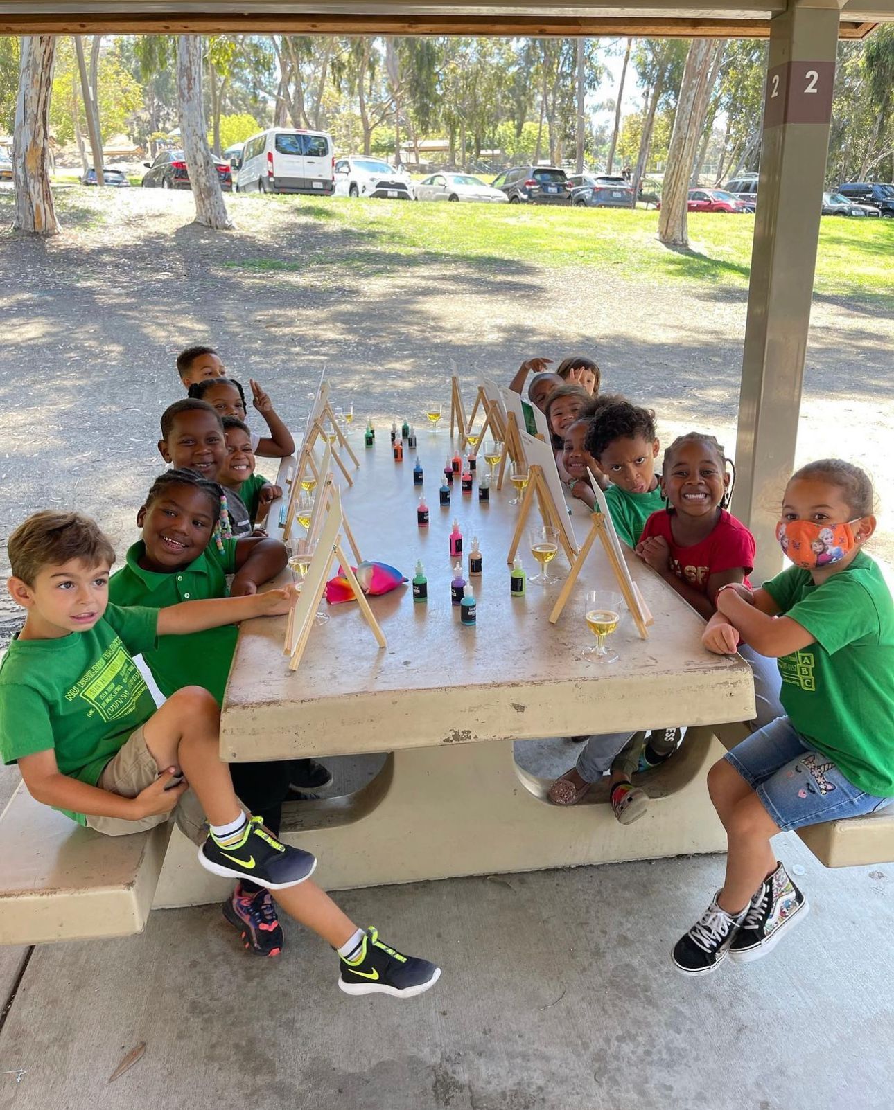 Children in green shirts at a picnic table with art supplies. They are smiling and looking at the camera in a park setting.