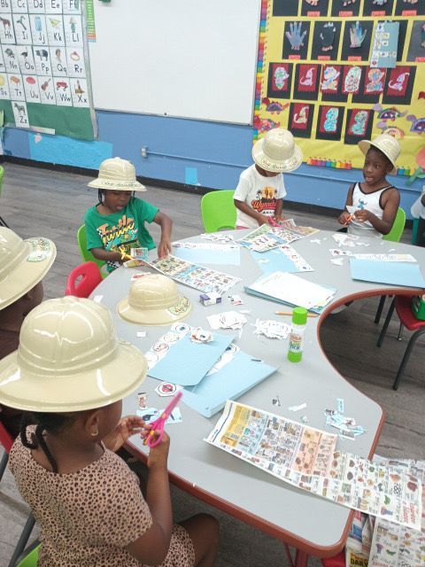 Children in safari hats at a table, crafting. Stickers, paper, glue, and colorful walls are in the classroom.