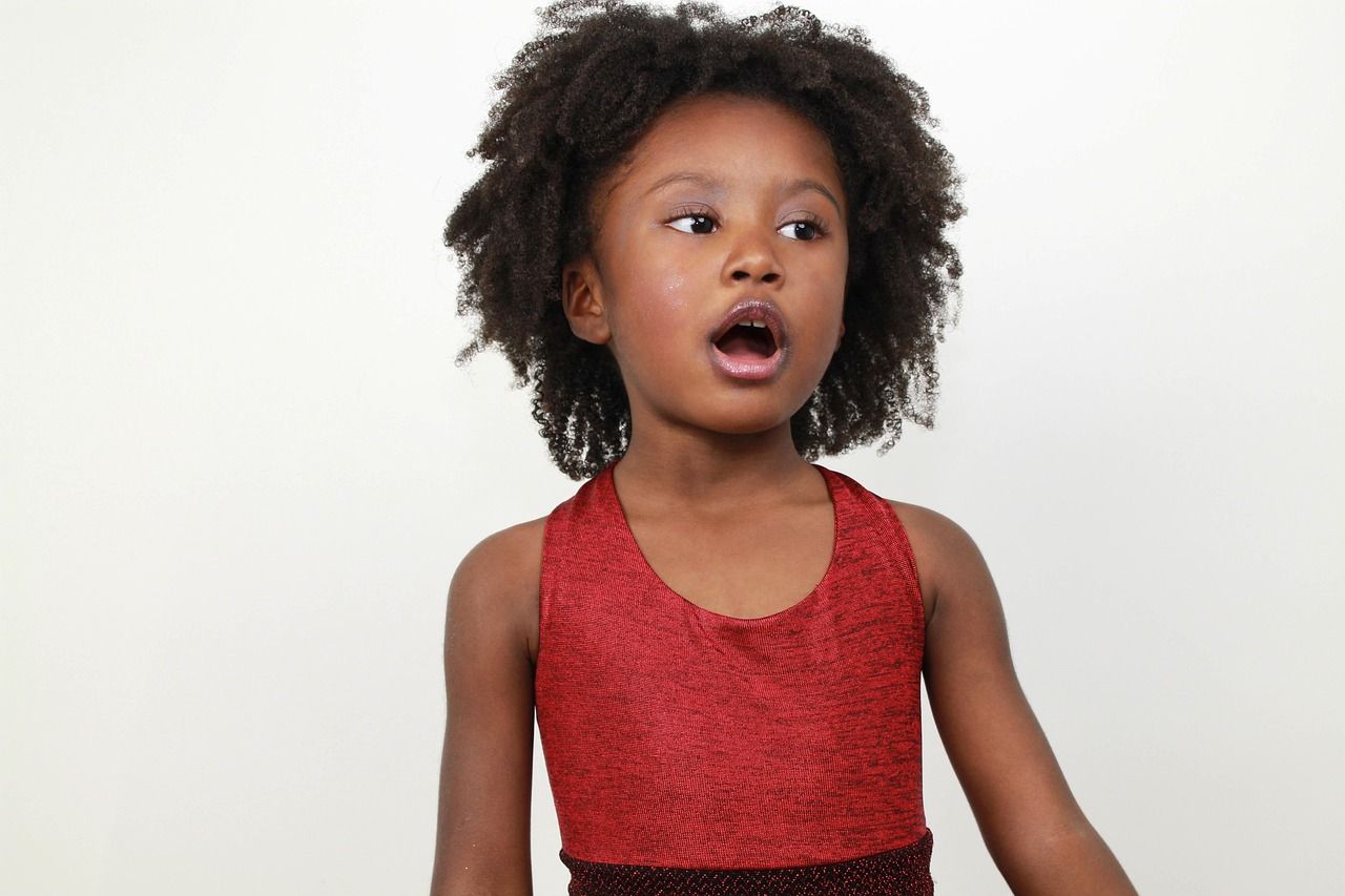 Young Black girl with curly hair, wearing a sparkly red dress, mouth open in surprise, looking to the side.