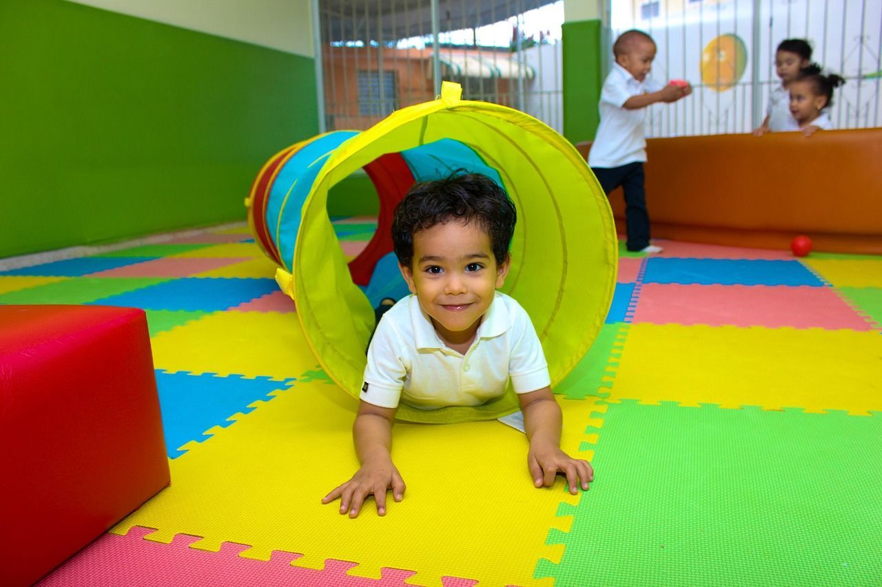A young boy crawls out of a colorful play tunnel in a brightly colored playroom. Other children play nearby.