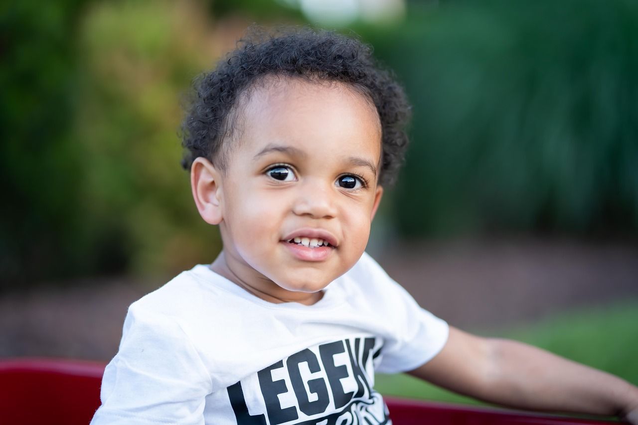 A young Black child with curly hair and a white shirt smiles, sitting outside in a red wagon.