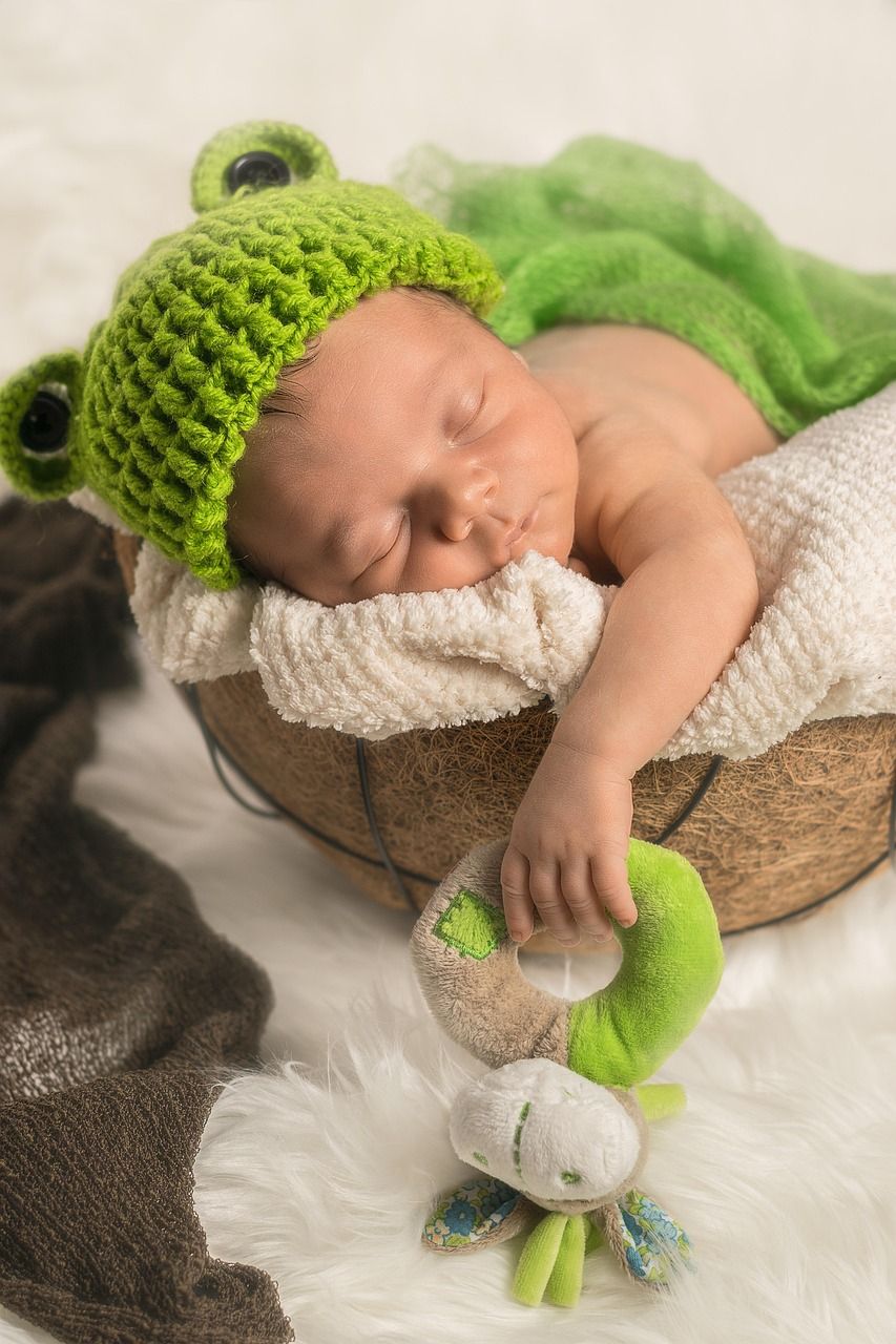 Newborn baby wearing a green frog hat, asleep in a basket, holding a green toy.