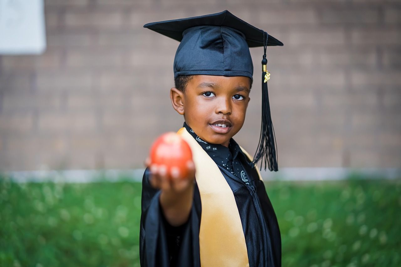 Young Black child in graduation attire holding an apple, smiling at the camera outdoors.