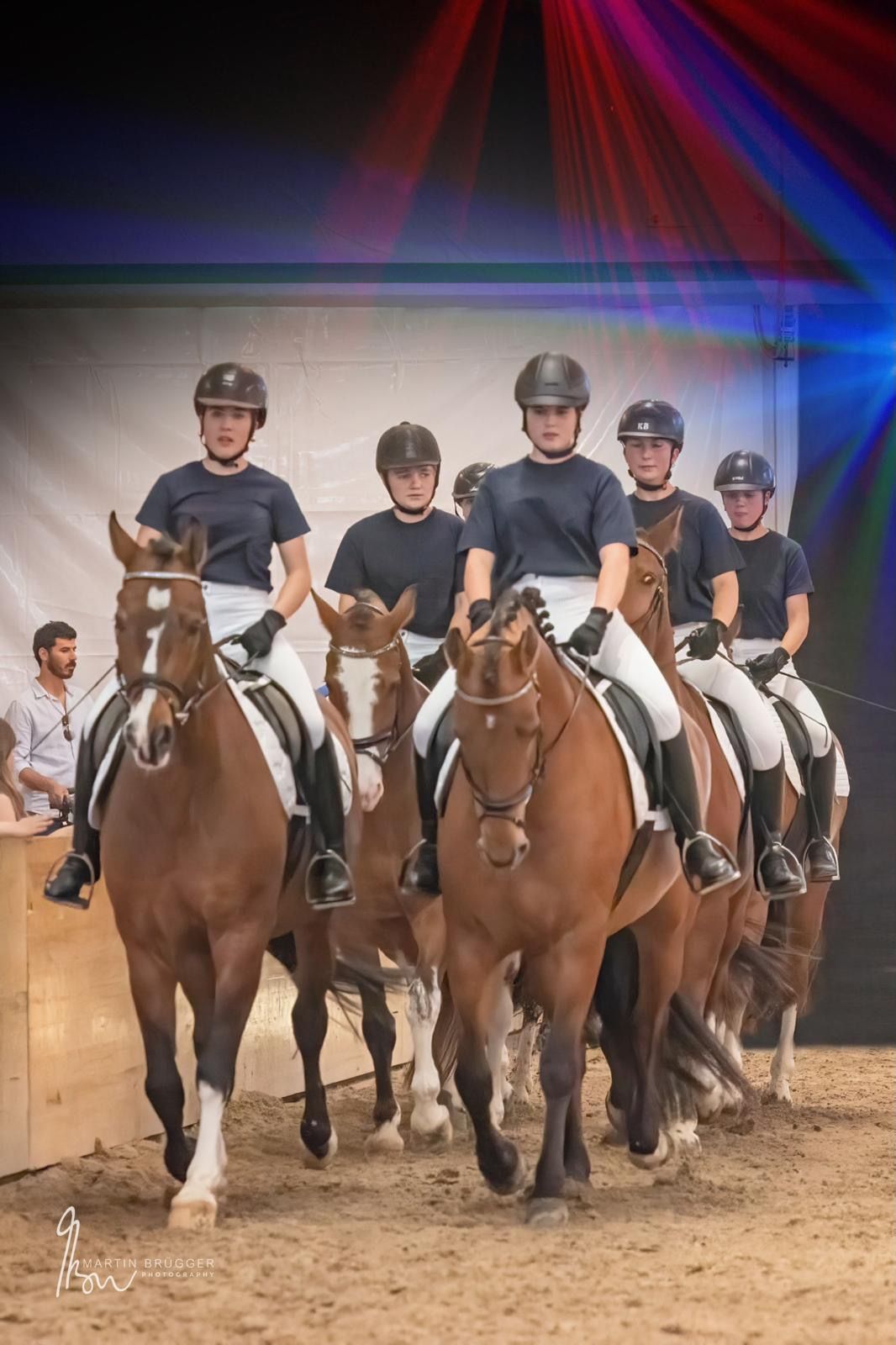 Sechs Reiter auf braunen Pferden in einem Ring, alle mit Helmen und dunkelblauen Hemden.