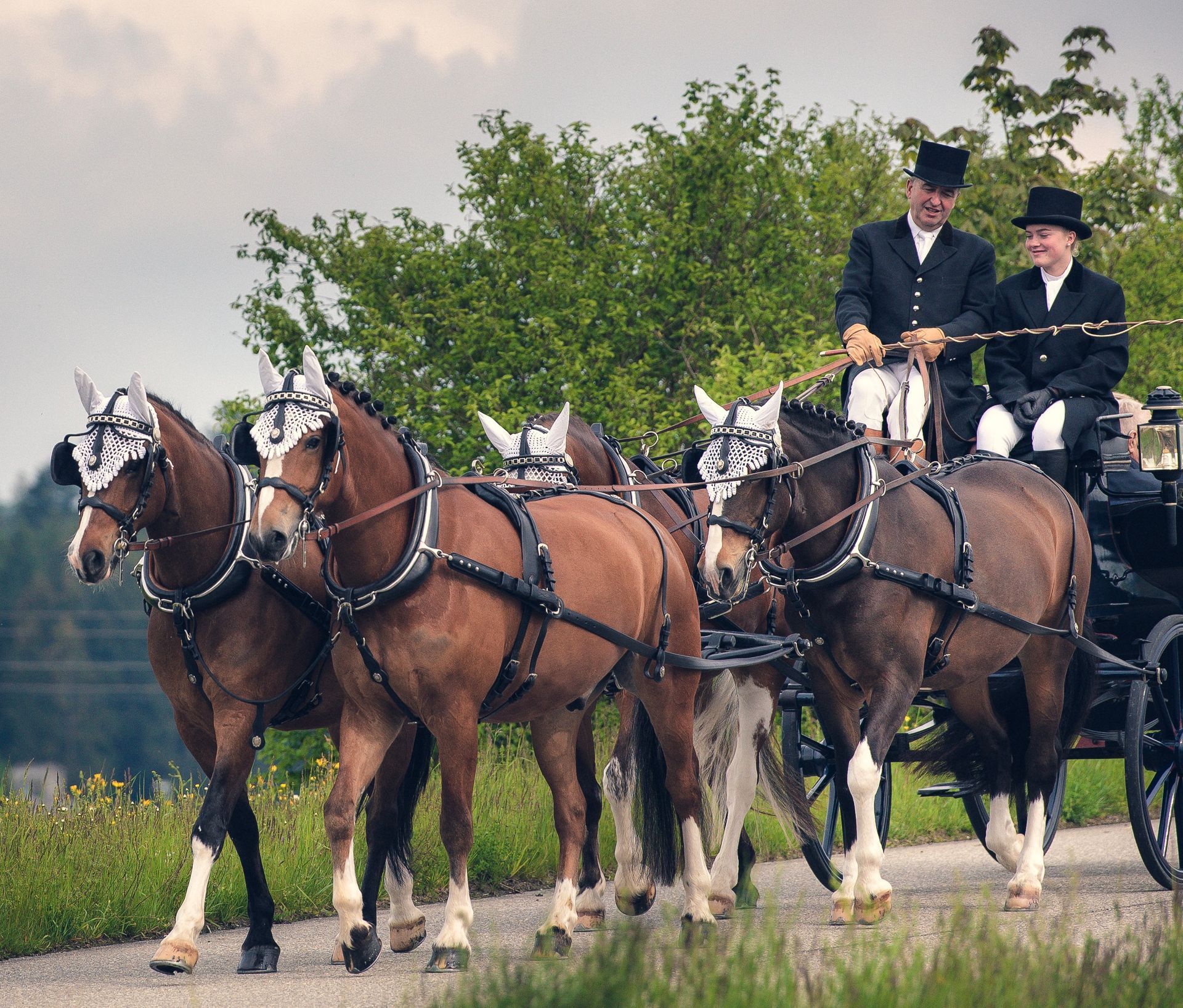 Vier braune Pferde ziehen eine Kutsche mit zwei Personen in Zylinderhüten auf einer Straße.