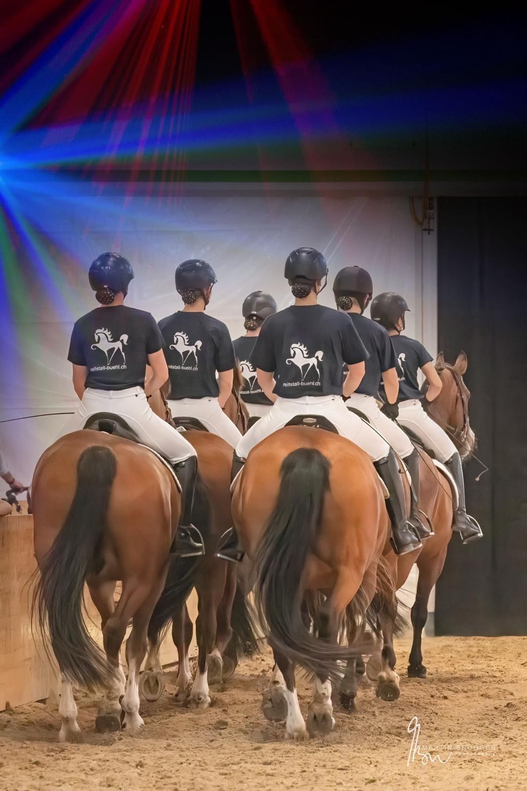 Eine Gruppe Reiter auf braunen Pferden, die den Rücken zugewandt haben. Sie tragen Helme, schwarze Hemden und weiße Hosen und reiten in einer Arena.
