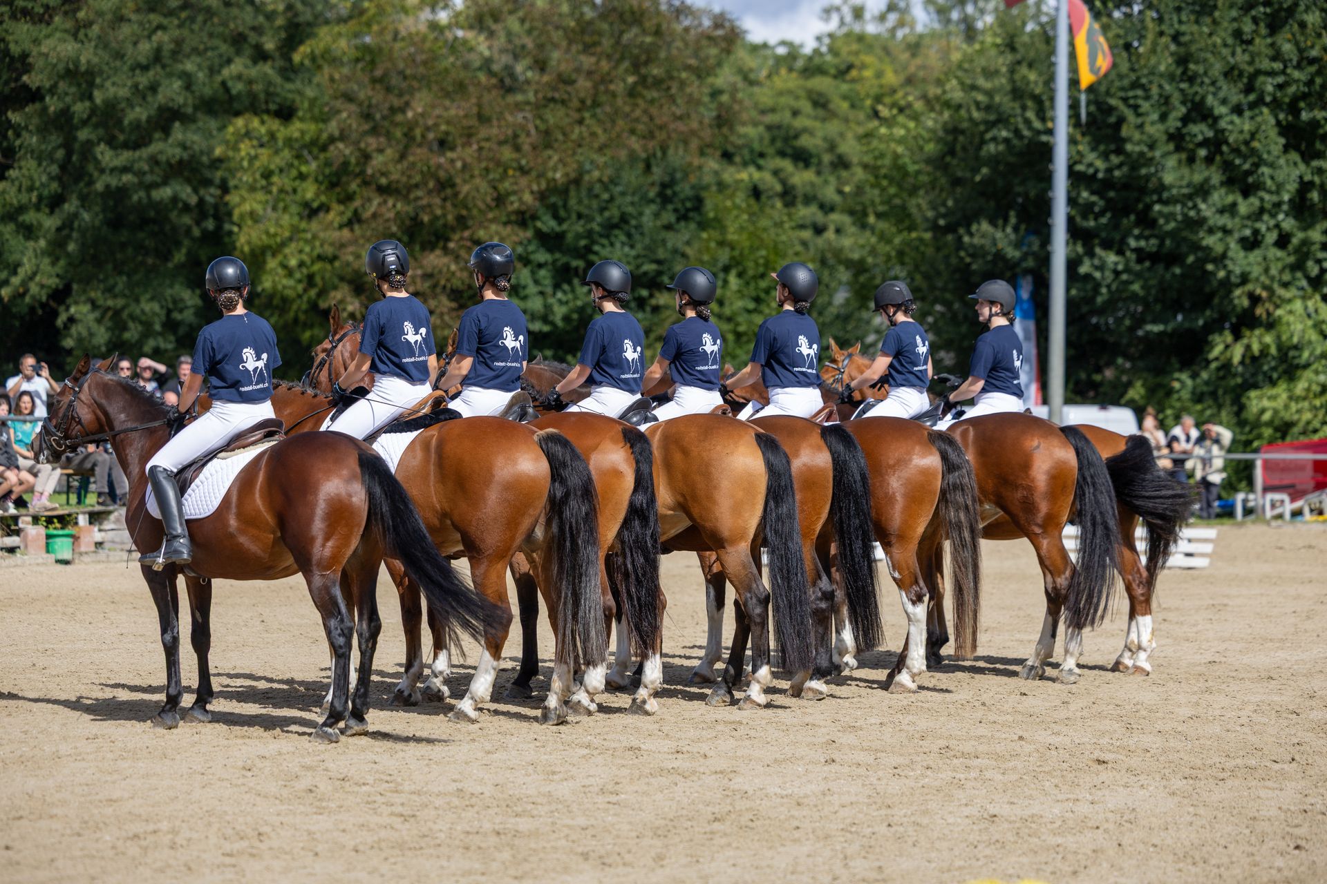 Eine Reihe von Reitern zu Pferd, die einheitliche marineblaue Oberteile und Helme tragen, in einer Reitanlage im Freien.