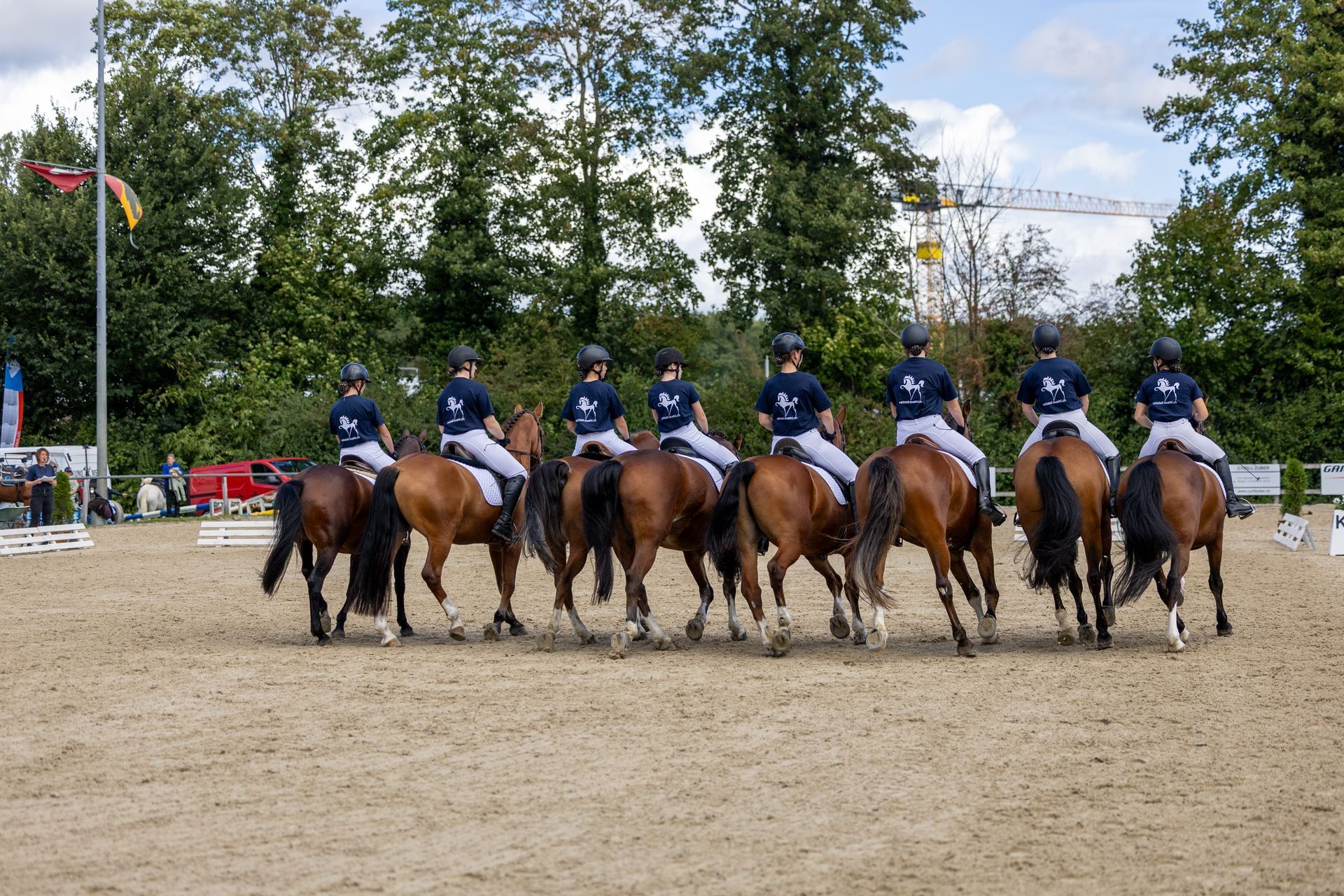 Acht Reiter treten zu Pferd in einer sandigen Arena auf, im Hintergrund sind Bäume zu sehen.