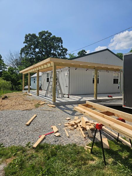 Construction of a wooden carport attached to a white building. Lumber, tools, and a ladder are present.