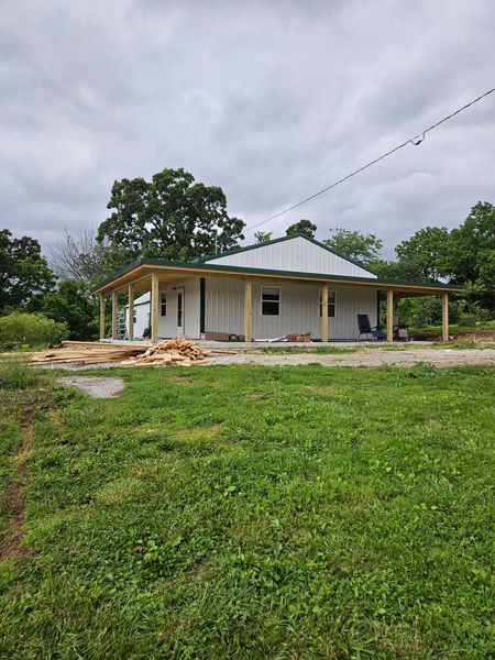 A partially constructed white house with a green roof and wooden porch, in a grassy field.