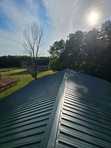 Green metal roof with concentric lines, sunlight, and a view of trees and fields.