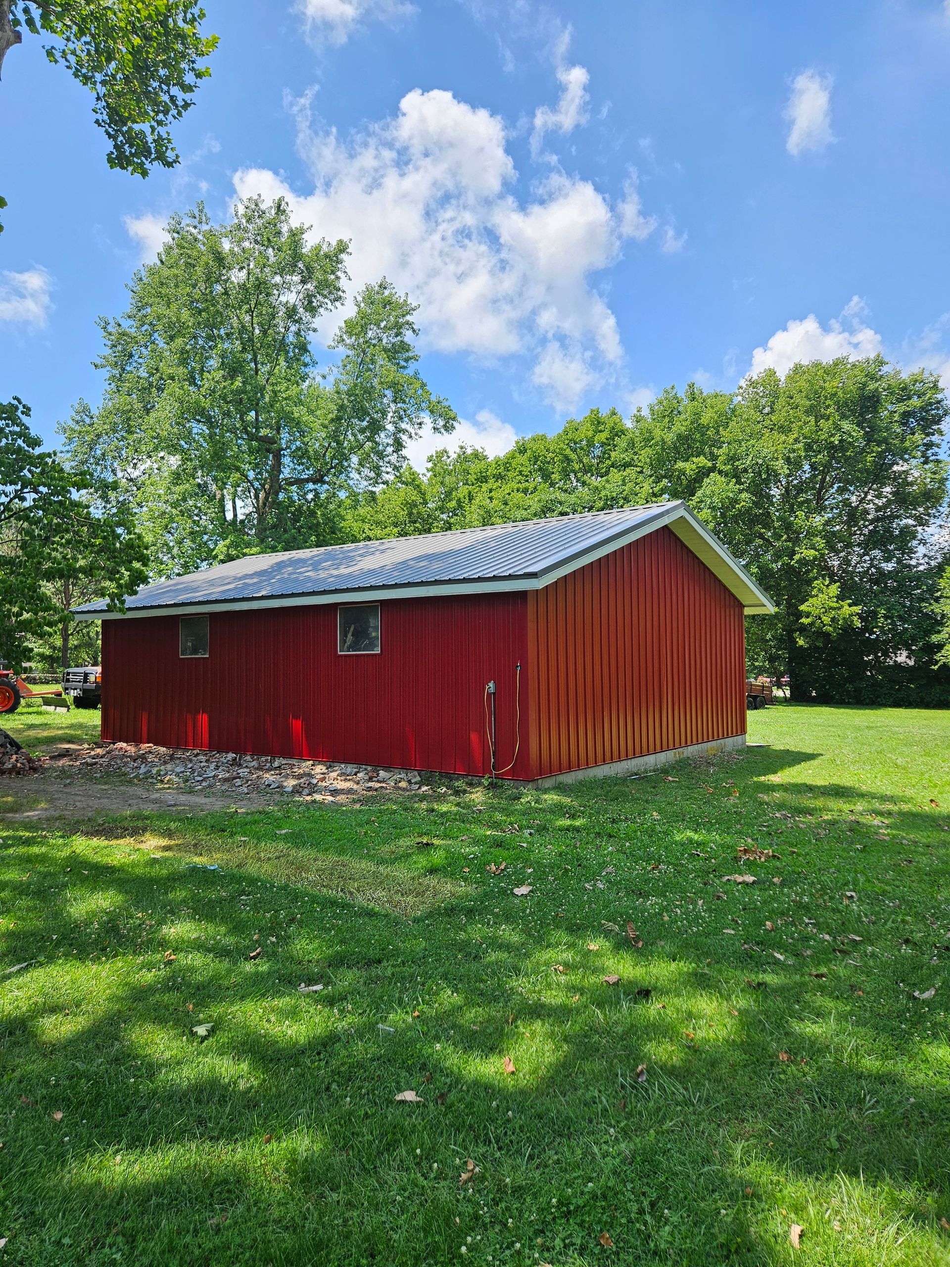 Red barn with a dark roof on a grassy lawn under a blue sky with trees.