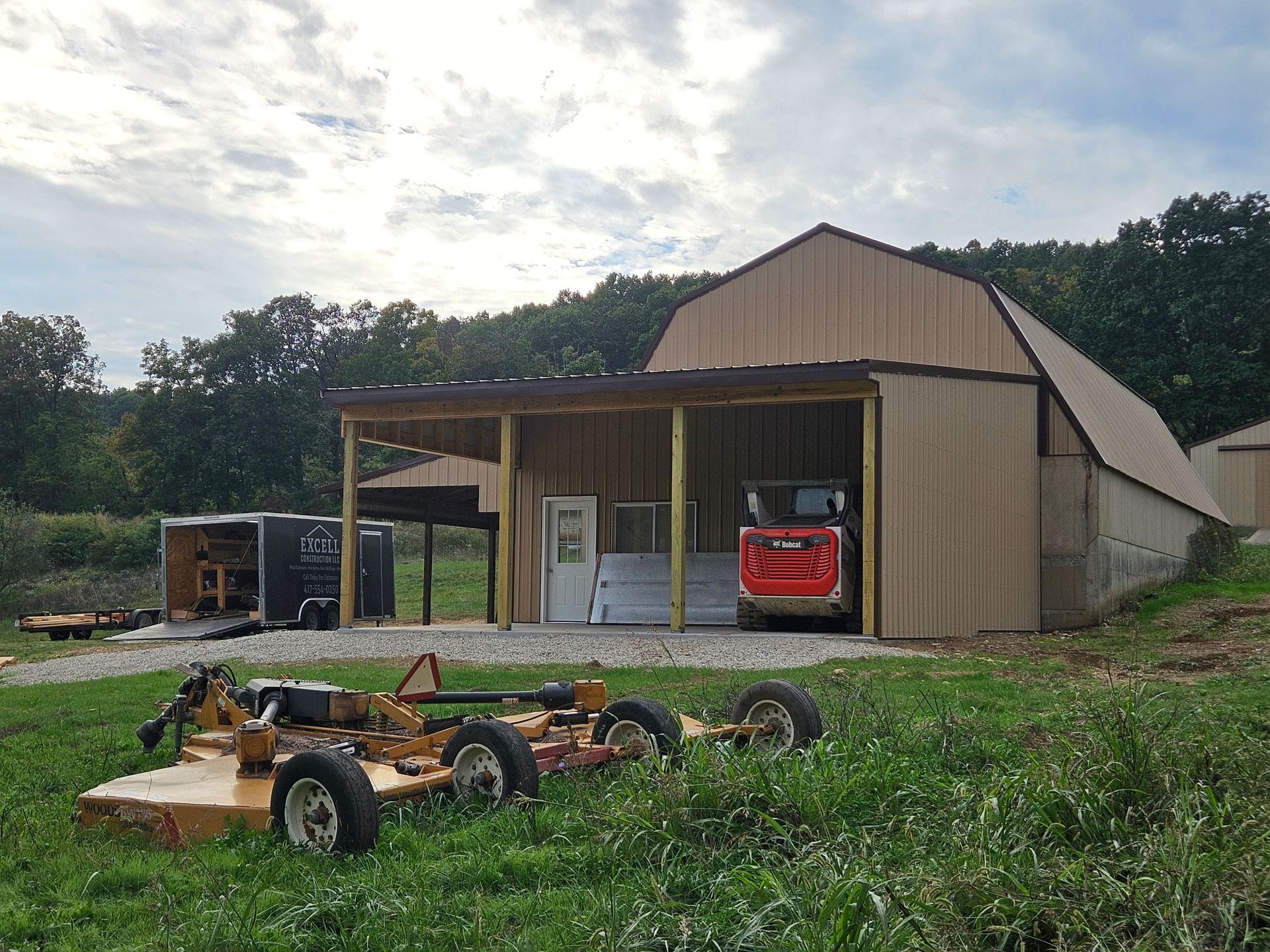 Barn with an open bay holding a red Bobcat. A mower sits in the foreground, with trees and a cloudy sky in the background.