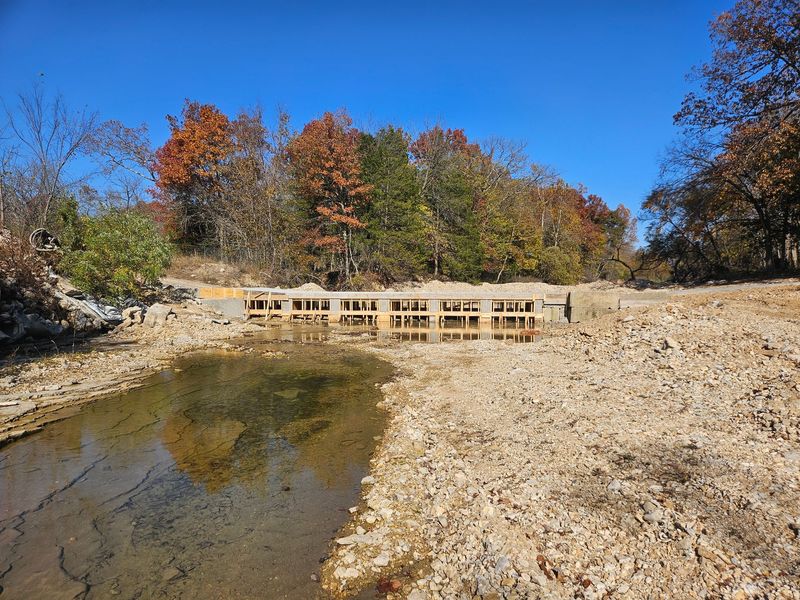 A wooden barrier in a dried-up riverbed, autumn foliage in the background under a blue sky.