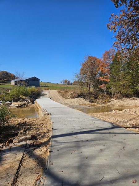 Concrete path leads uphill to a distant gray building under a clear blue sky, with trees on the right and dirt edges.