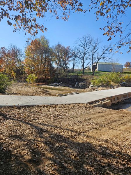 A paved pathway crosses a rocky creek bed, surrounded by trees with fall foliage under a clear blue sky.